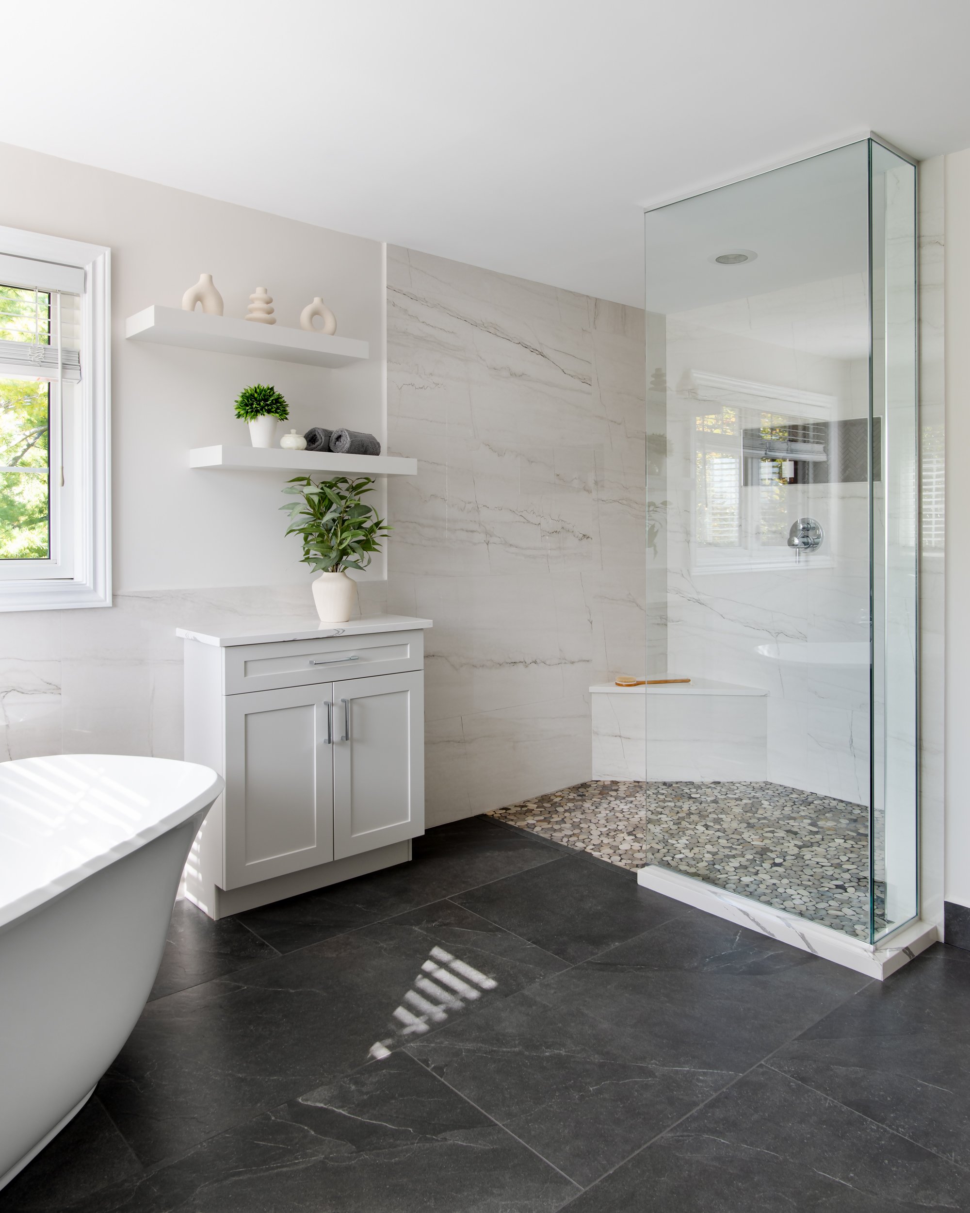 Modern bathroom with a freestanding bathtub, a glass-enclosed walk-in shower, white cabinetry, floating shelves with decor, a window with blinds, and dark tiled flooring.