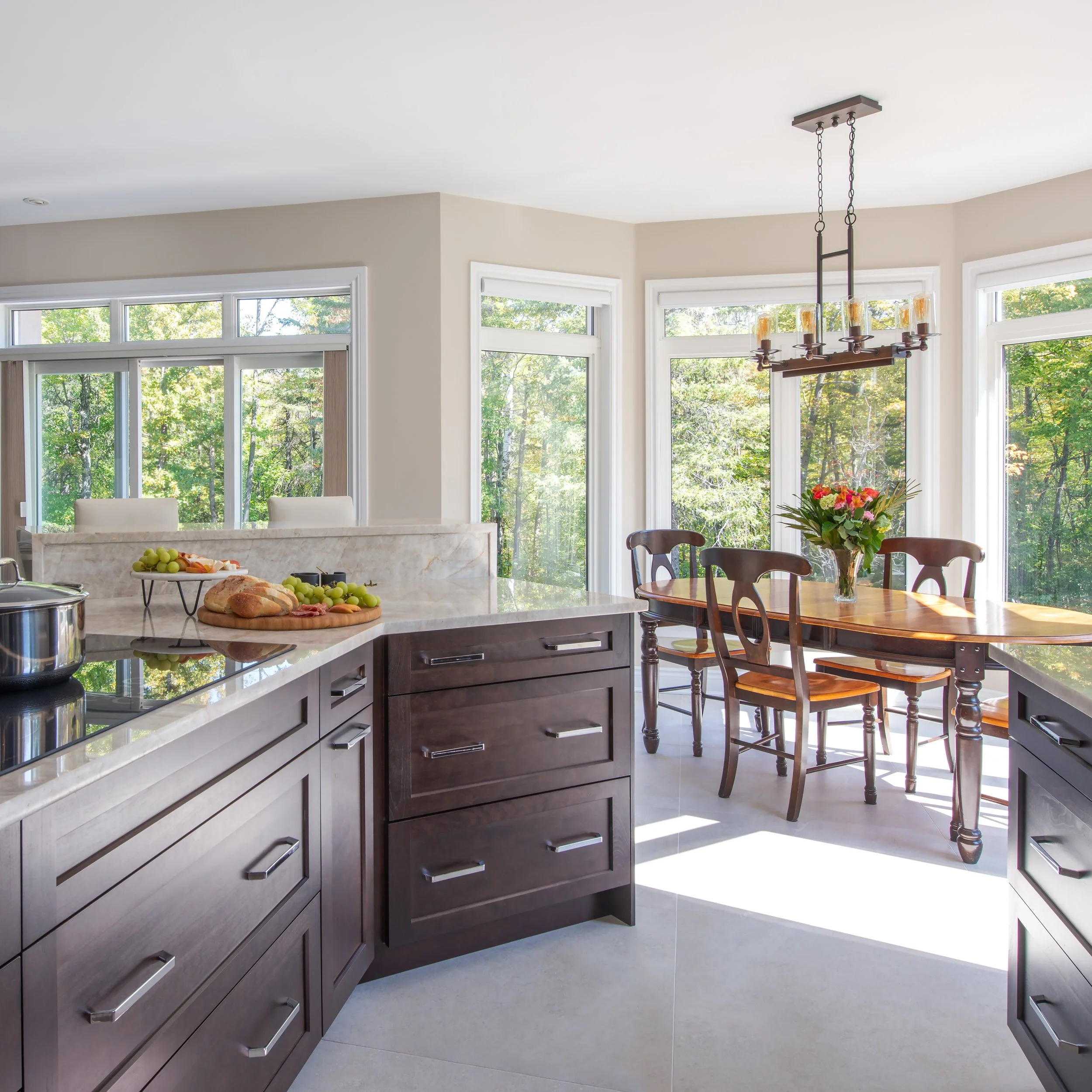 Bright kitchen with dark wood cabinets, a marble countertop, and a dining area with a wooden table, four chairs, and a hanging chandelier, surrounded by large windows showing green trees outside.