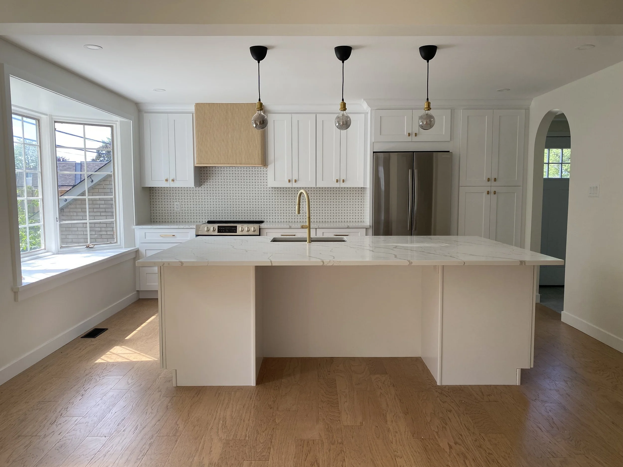 White bathtub with brushed nickel faucet and handles, located in a bathroom with gray tiled wall and a window with white trim, with a green plant in a black metal basket nearby.