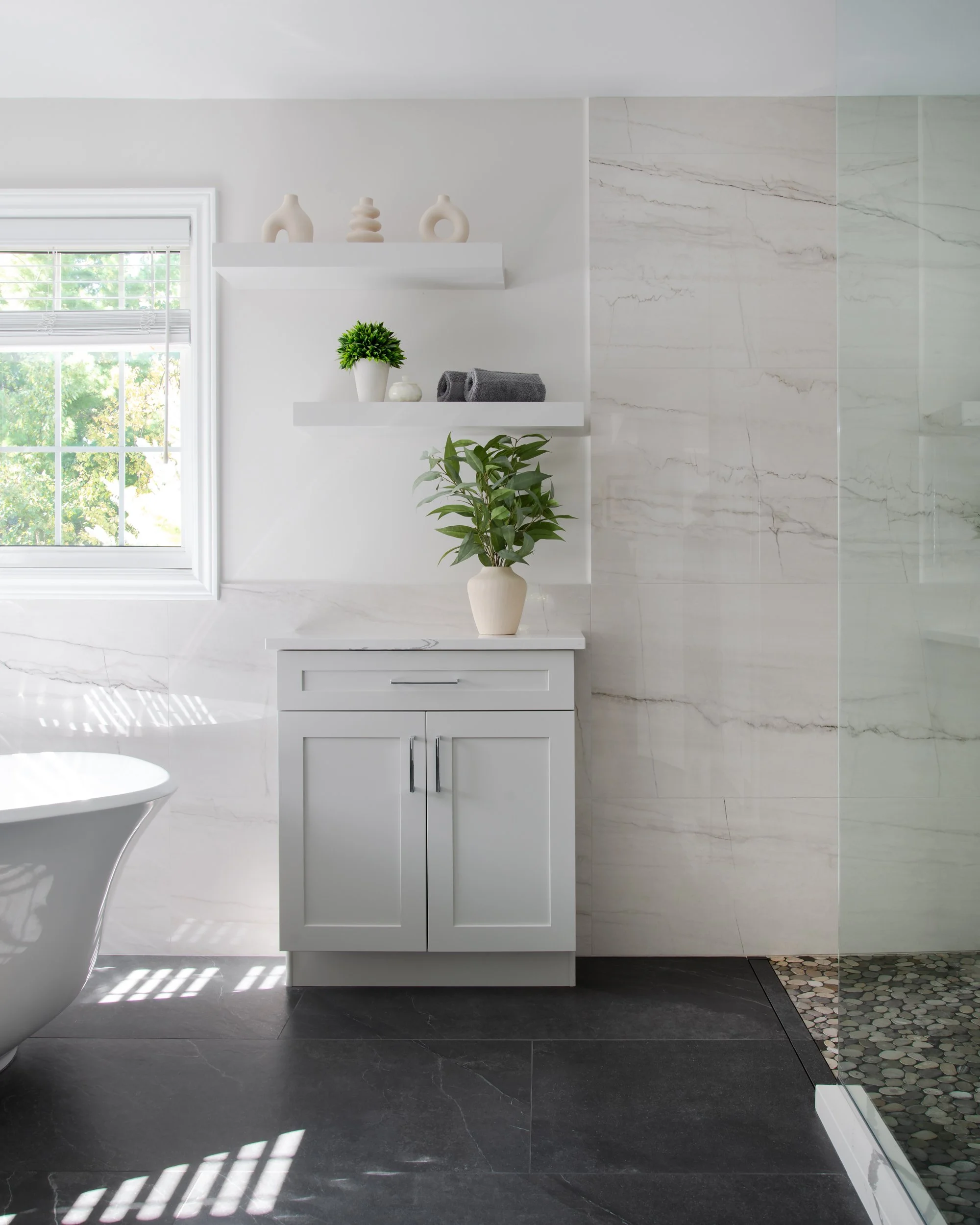 A modern bathroom with white walls, a window with blinds, and decorated with vases, a potted plant on a white cabinet, hanging shelves with decor items, and a section of a bathtub.