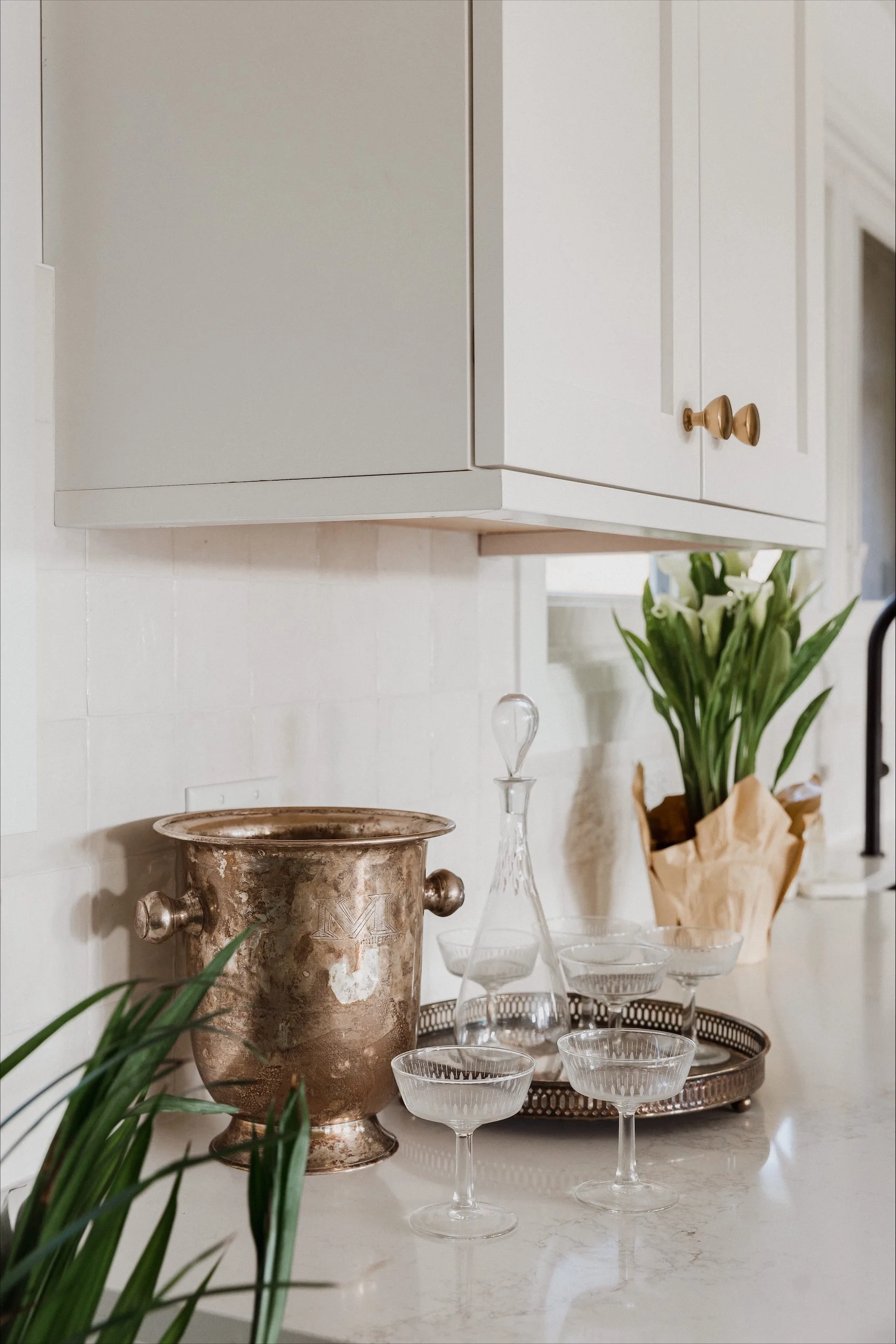 Modern bathroom with light-colored walls and flooring, featuring a large mirror above a white vanity with gold hardware, a bathtub with a glass partition, and a ladder towel rack near a window, decorated with minimalistic art and decor.