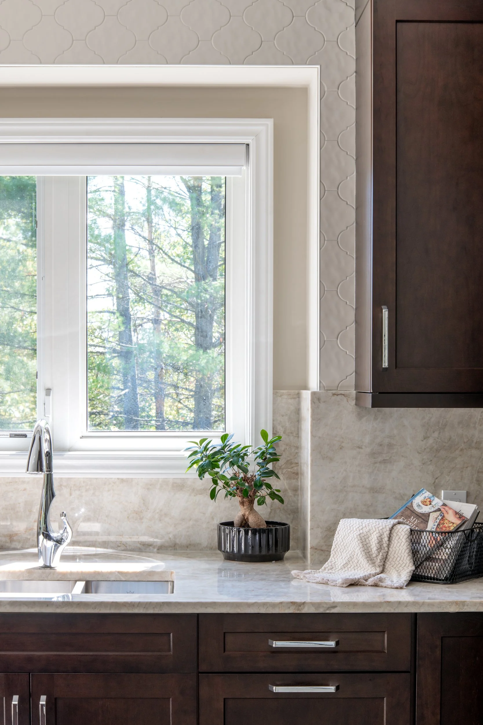 Kitchen window with a view of trees, a potted bonsai plant, and a basket with magazines and a cloth on the countertop.