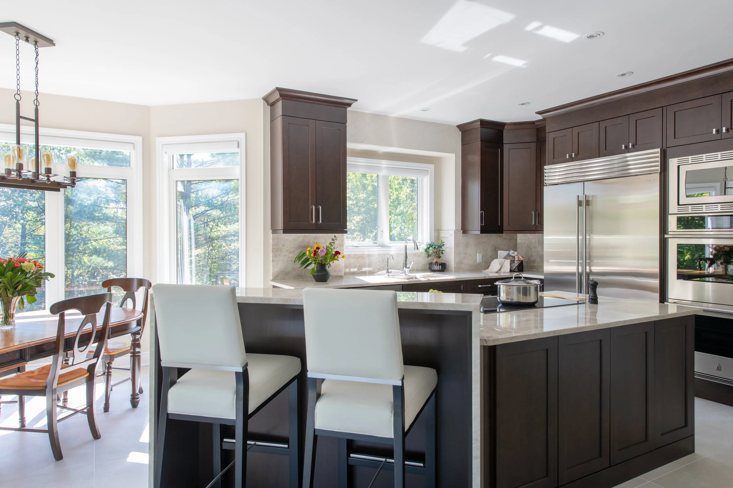 Modern kitchen with dark wood cabinets, stainless steel appliances, marble countertops, and large windows with outdoor greenery.