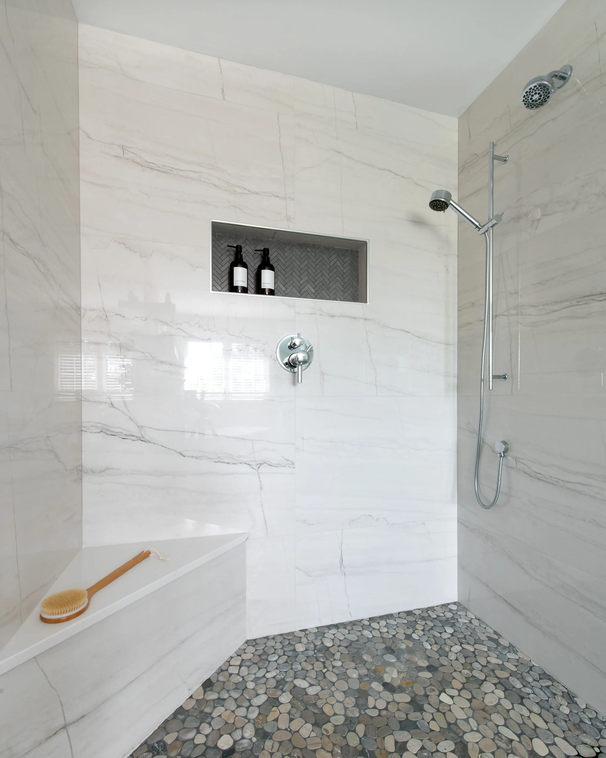 Modern shower area with marble walls, pebble stone floor, built-in niche with black bottles, and a handheld showerhead.