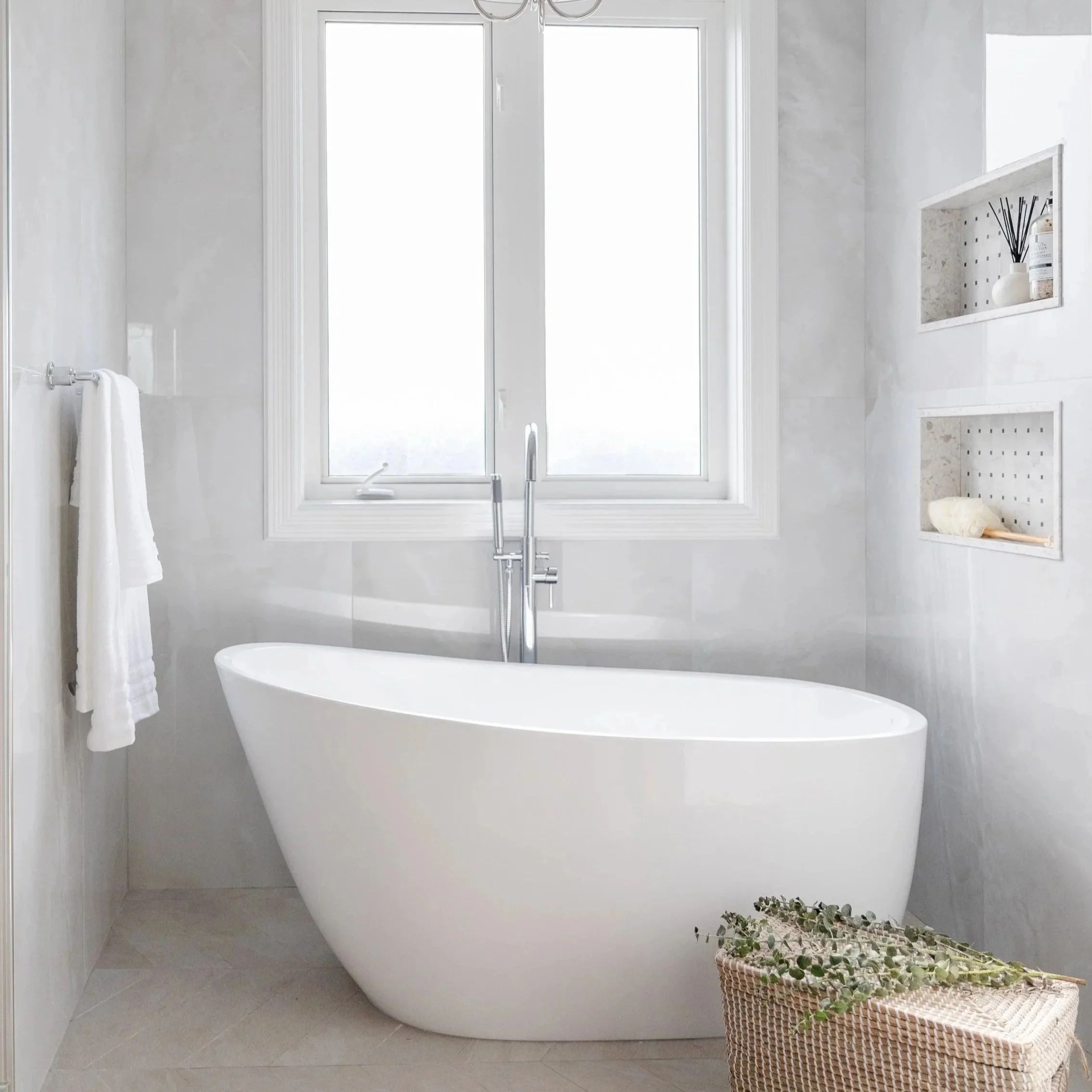 Modern white bathtub in a bright bathroom with large window, neutral wall color, built-in wall niches, and a woven basket with greenery.