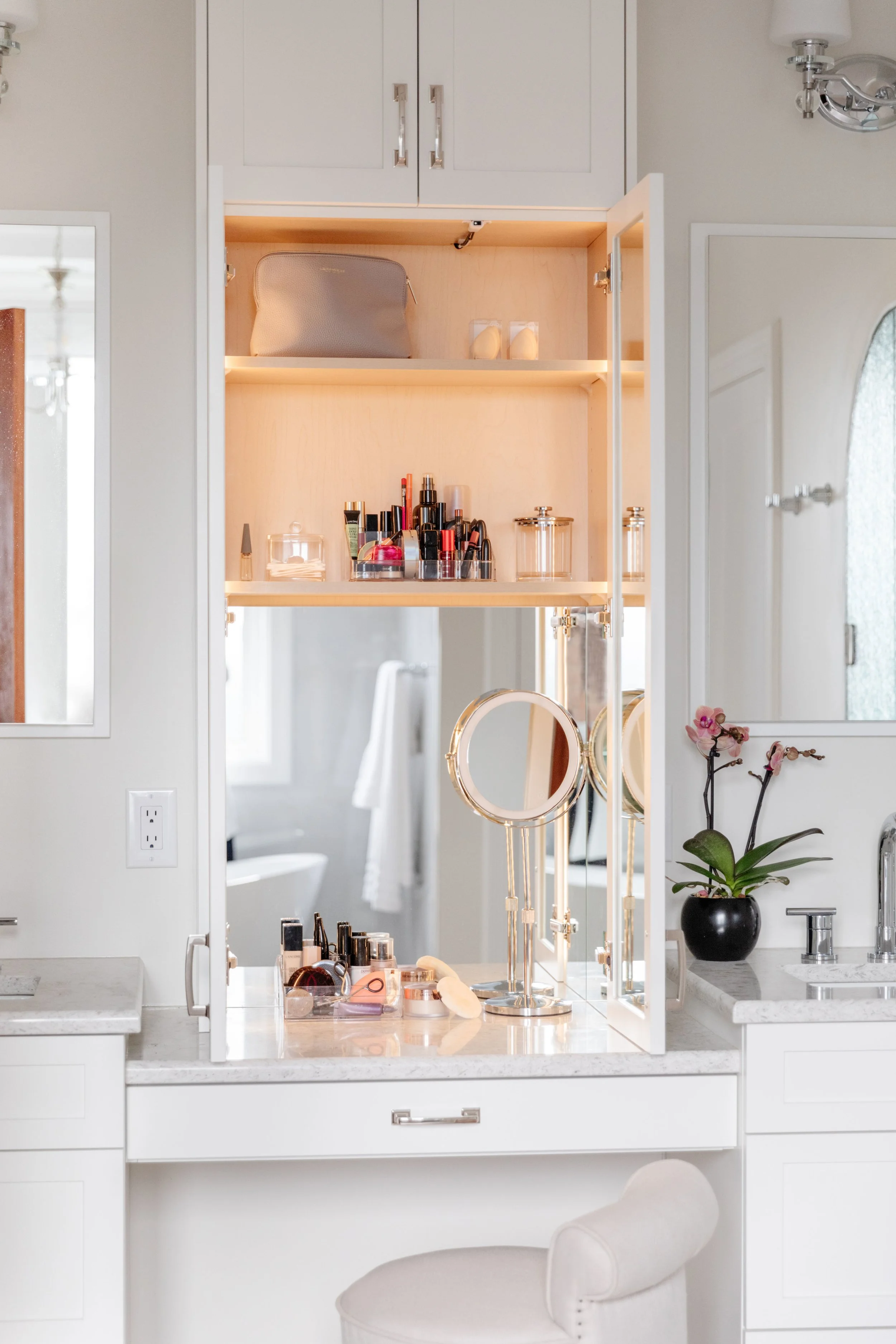 A bathroom vanity with a large mirror, makeup and skincare products, a black orchid plant, and a cabinet with shelves holding various items. The background shows a bathroom with a bathtub and towel.