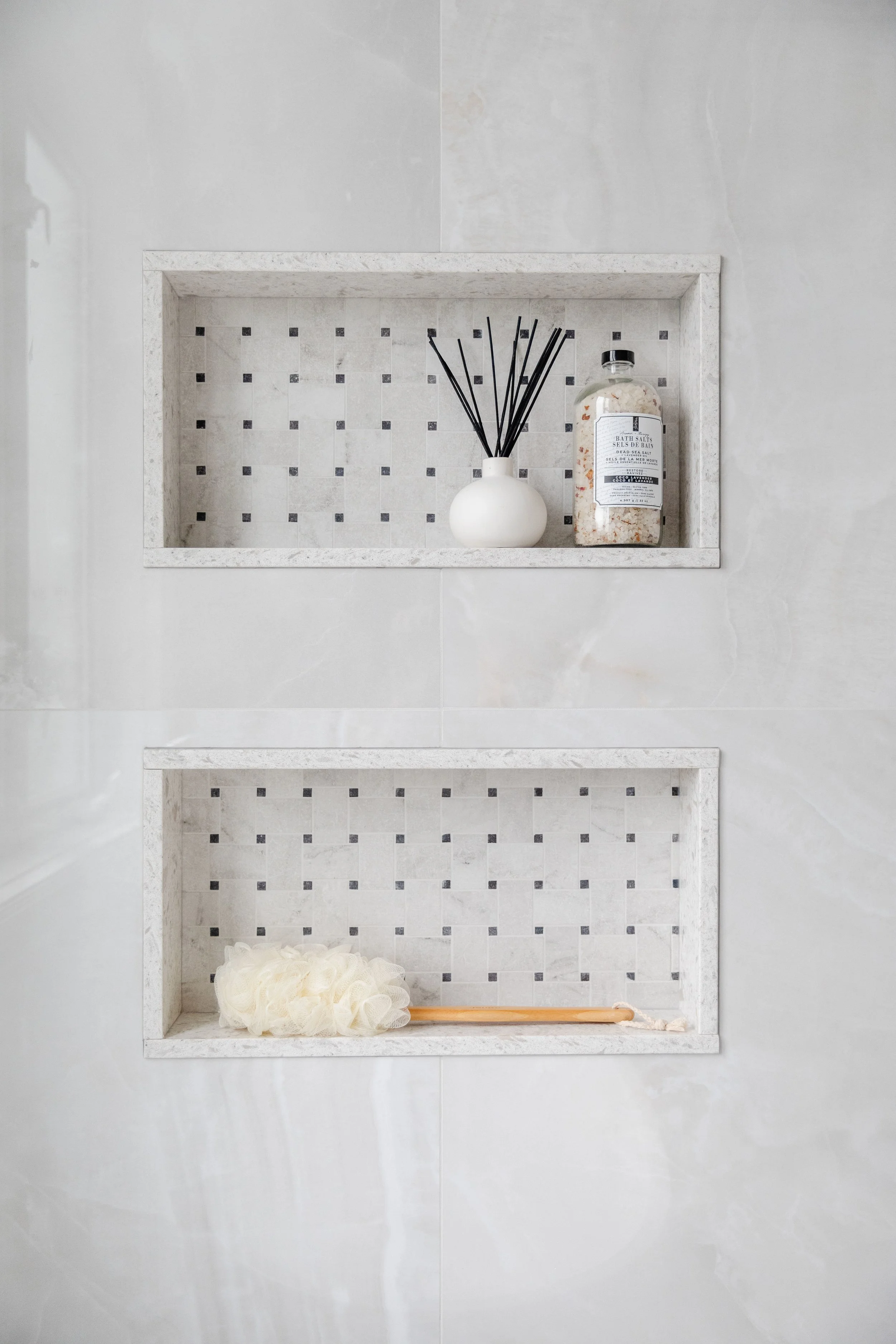 Two built-in shower shelves with a spa-like decor. The top shelf holds a white vase with black reed diffuser sticks and a bottle of bath salts. The bottom shelf has a bath loofah and a wooden bath brush.