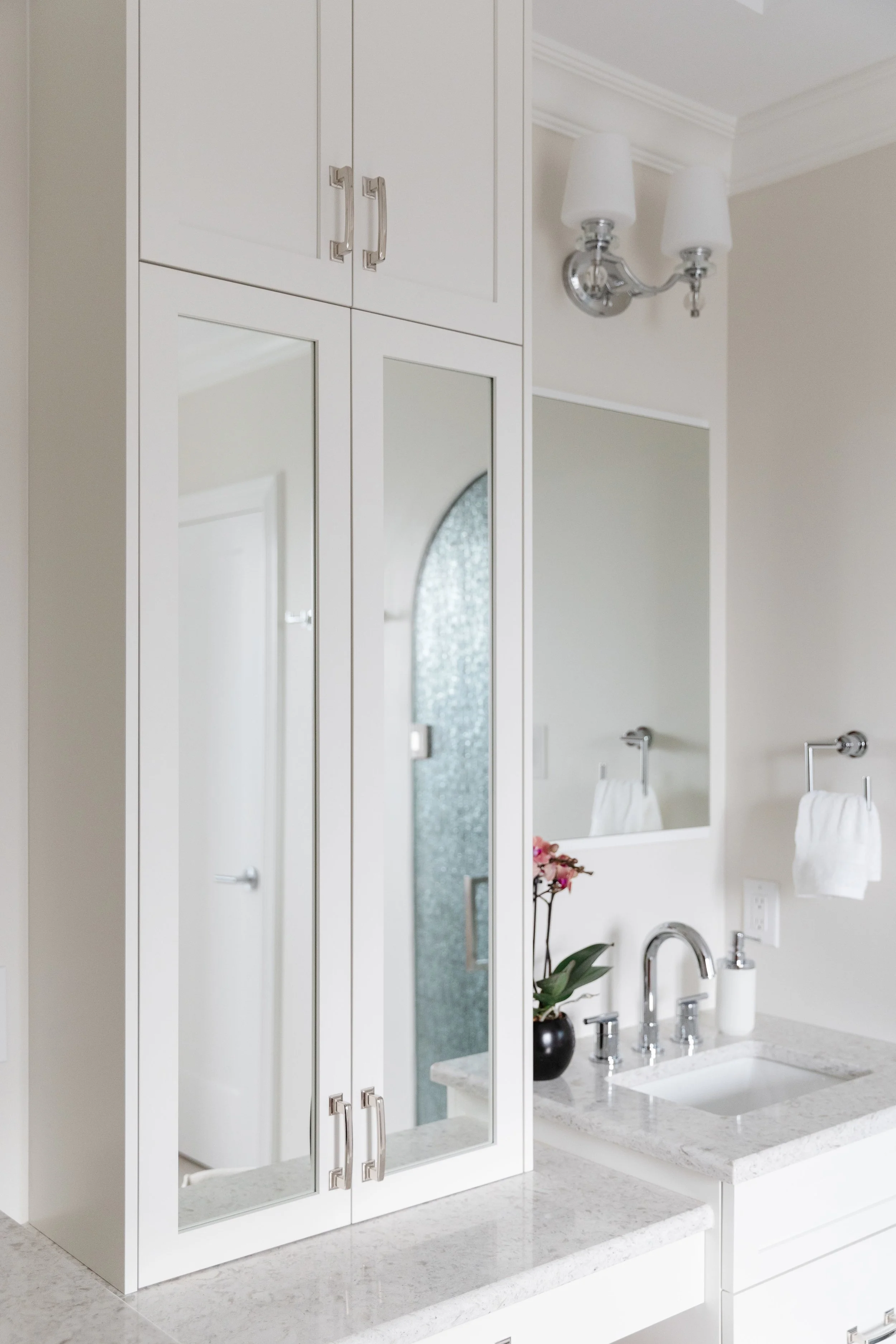 A white bathroom vanity with a granite countertop, a small black vase with pink flowers, a wall-mounted mirror, a chrome faucet, white soap soap dispenser, two white towels on chrome towel bars, and a decorative wall light fixture.
