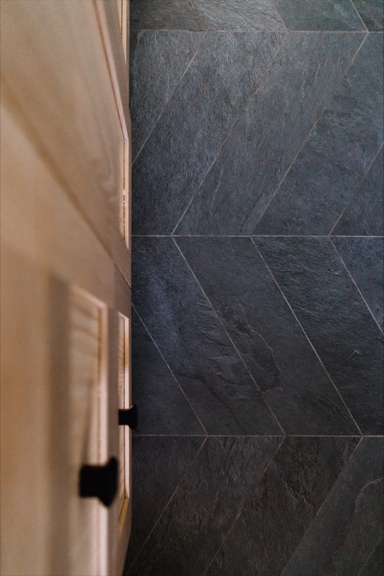 White bathtub with brushed nickel faucet and handles, located in a bathroom with gray tiled wall and a window with white trim, with a green plant in a black metal basket nearby.