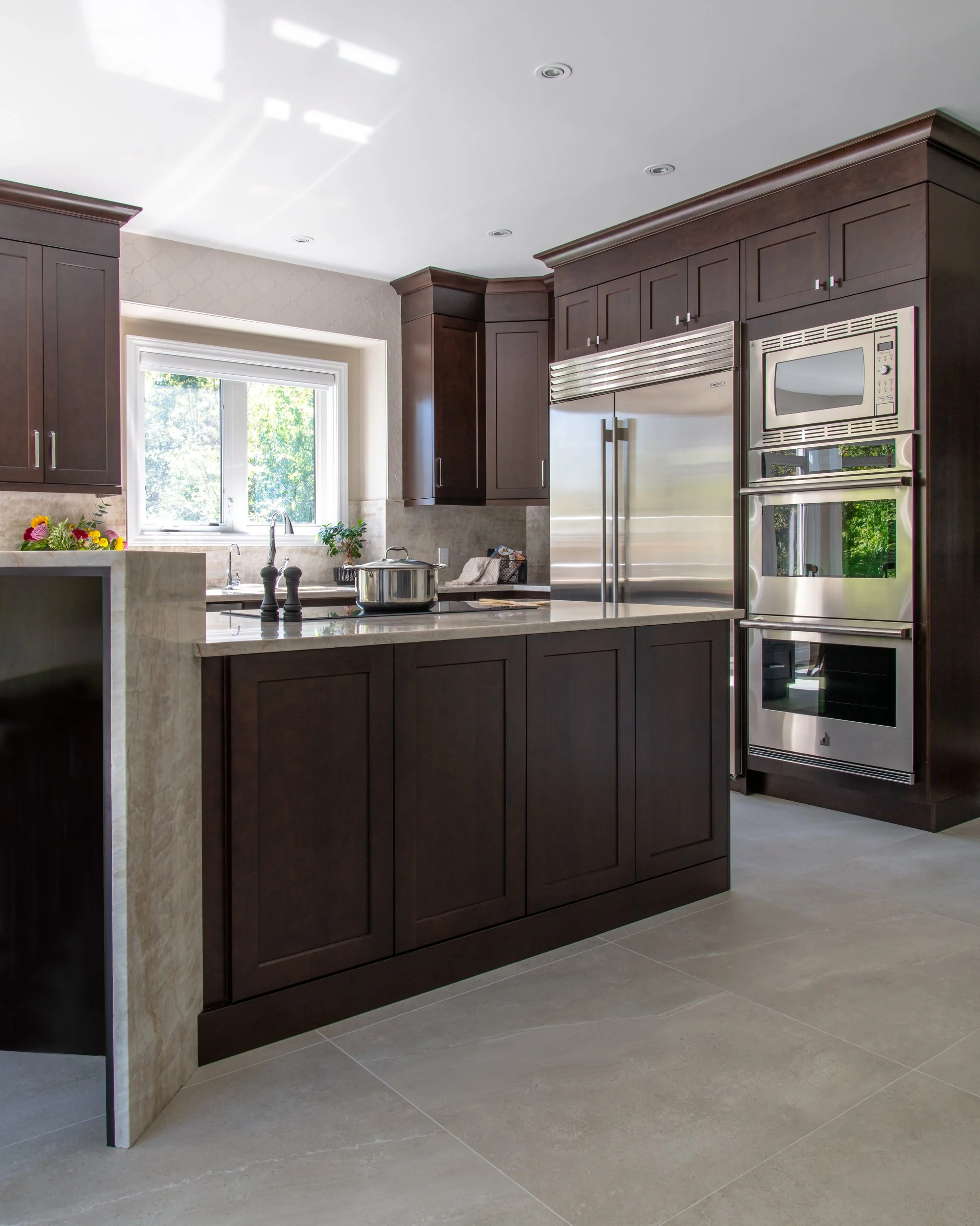 Modern kitchen with dark wood cabinets, stainless steel appliances, a window above the sink showing green trees outside, and a beige tiled floor.