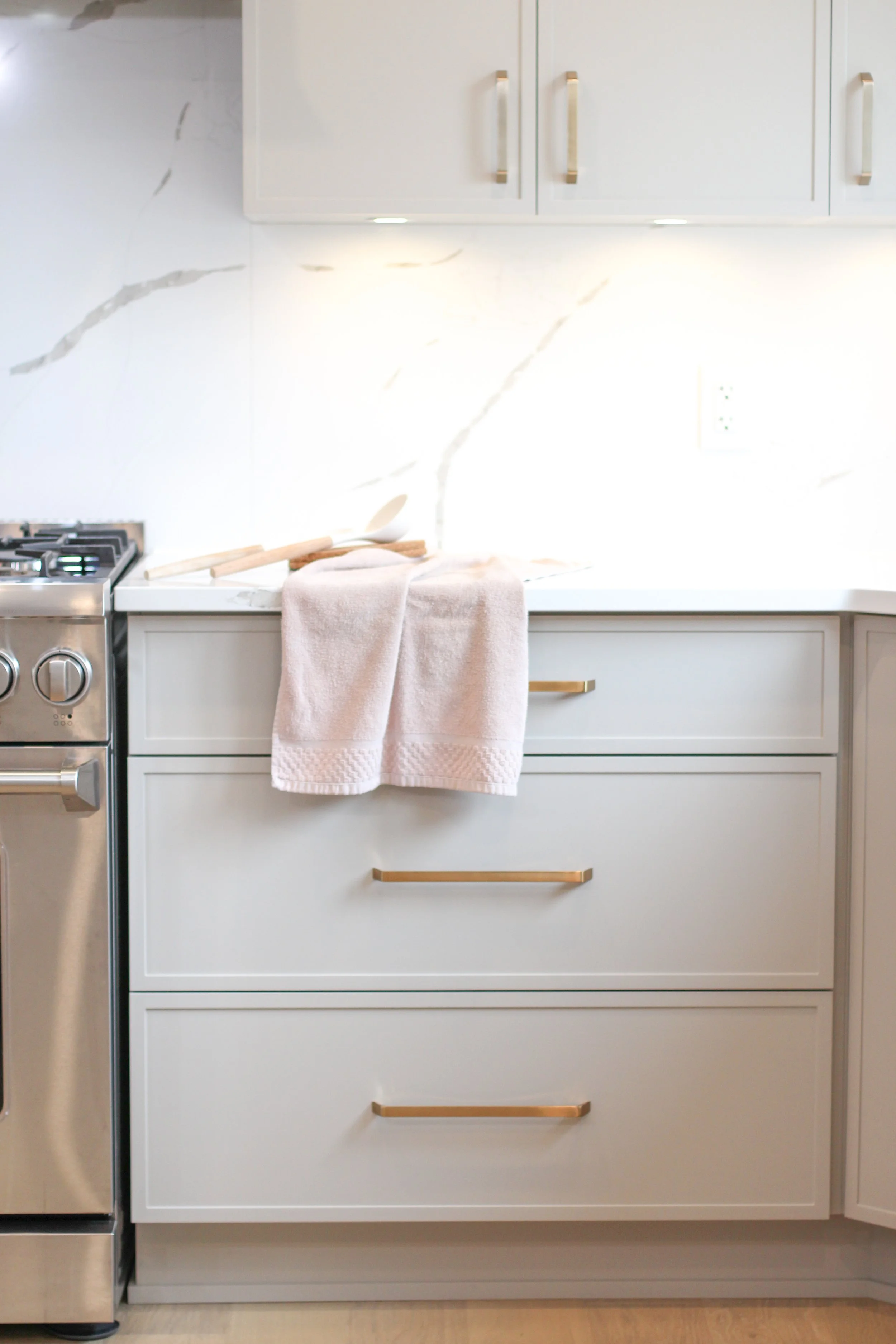 White kitchen cabinets with gold handles, a marble backsplash, a beige towel hanging over a drawer, and part of a stainless steel stove.