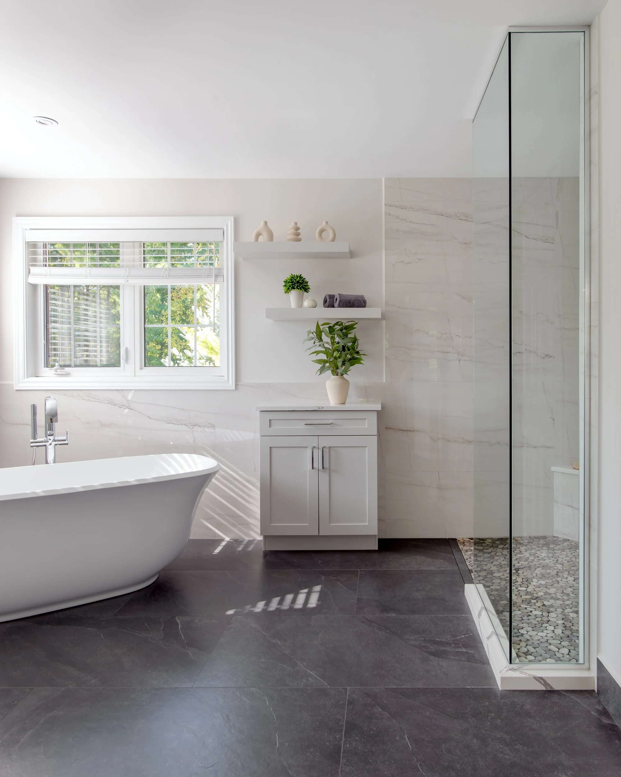 Modern bathroom with a bathtub, window with blinds, tiled shower, white cabinetry, and decorative vases and plant on shelves.