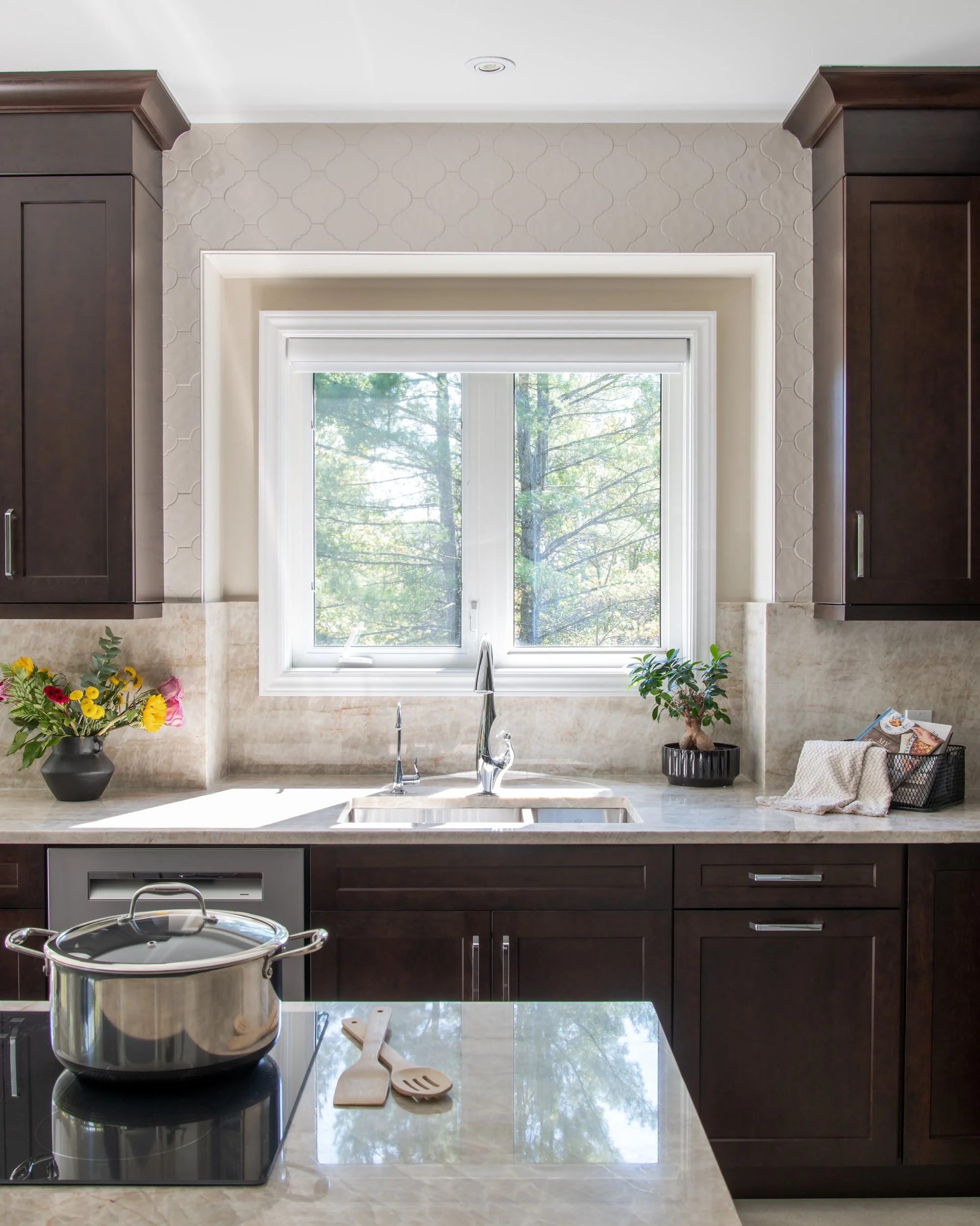 Modern kitchen with dark wood cabinets, beige marble countertops, and a window above the sink showing trees outside. There are flowers, a small plant, and kitchen utensils on the counter, and a pot on the stove.