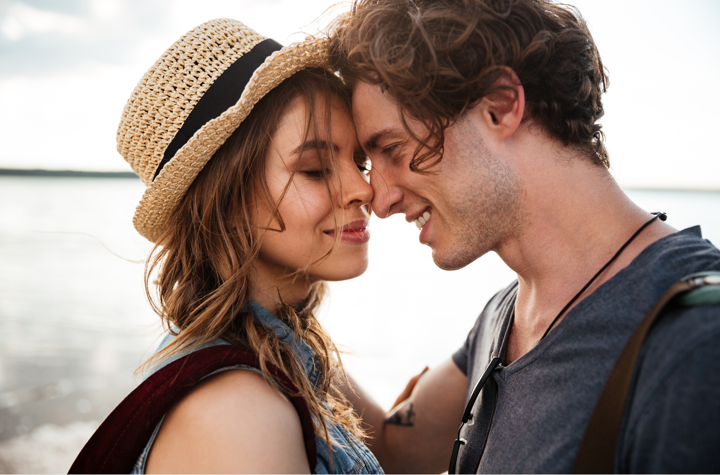 A couple standing close together with foreheads touching, both smiling, in front of water on a cloudy day. The woman wears a straw hat and a sleeveless top, and the man has curly hair and wears a dark t-shirt. They embrace in love.