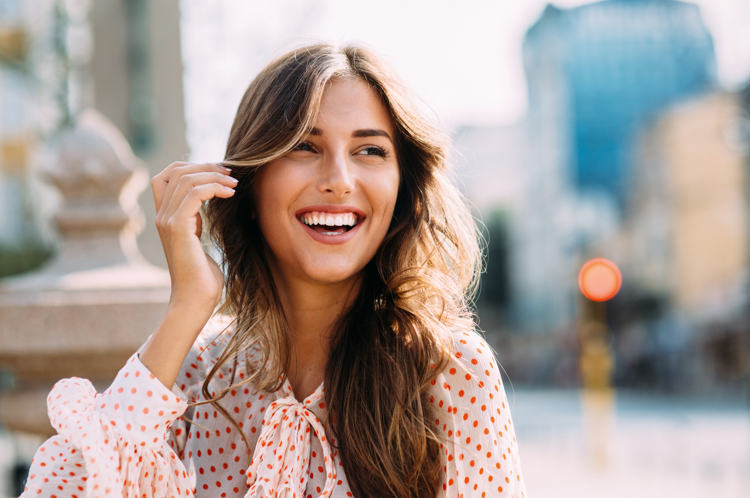Smiling woman with long hair in a polka dot blouse outdoors in a city setting
