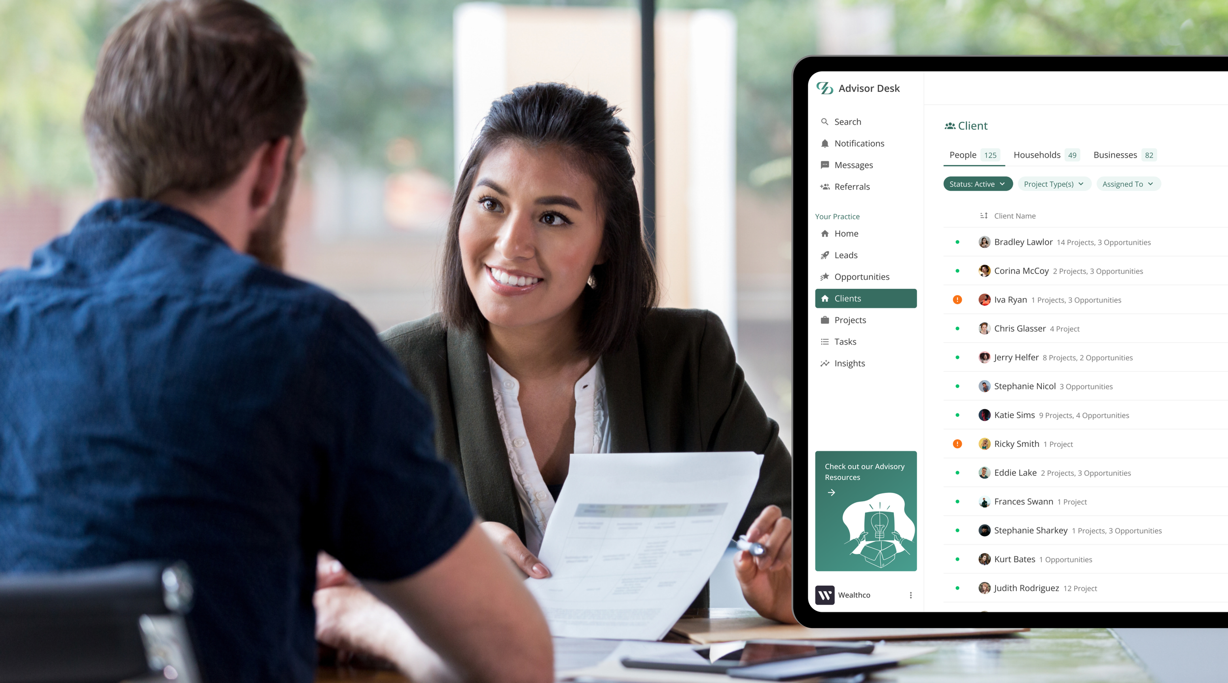 A woman sitting across from a man during a meeting, holding a document and smiling. In the background, a computer monitor displays a client management software dashboard with list of clients, projects, and opportunities.