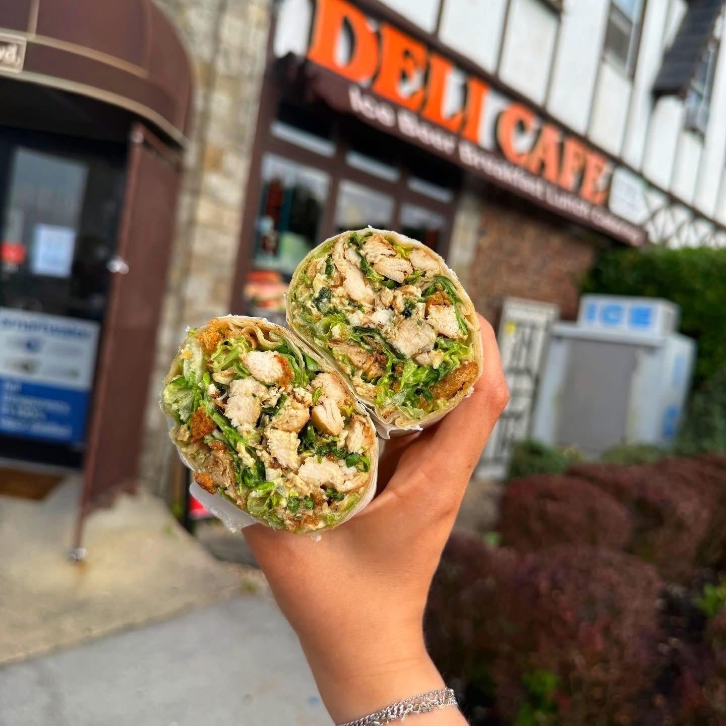 Hand holding a cut into a chicken salad wrap with lettuce, chicken, and vegetables outside a delicatessen store.