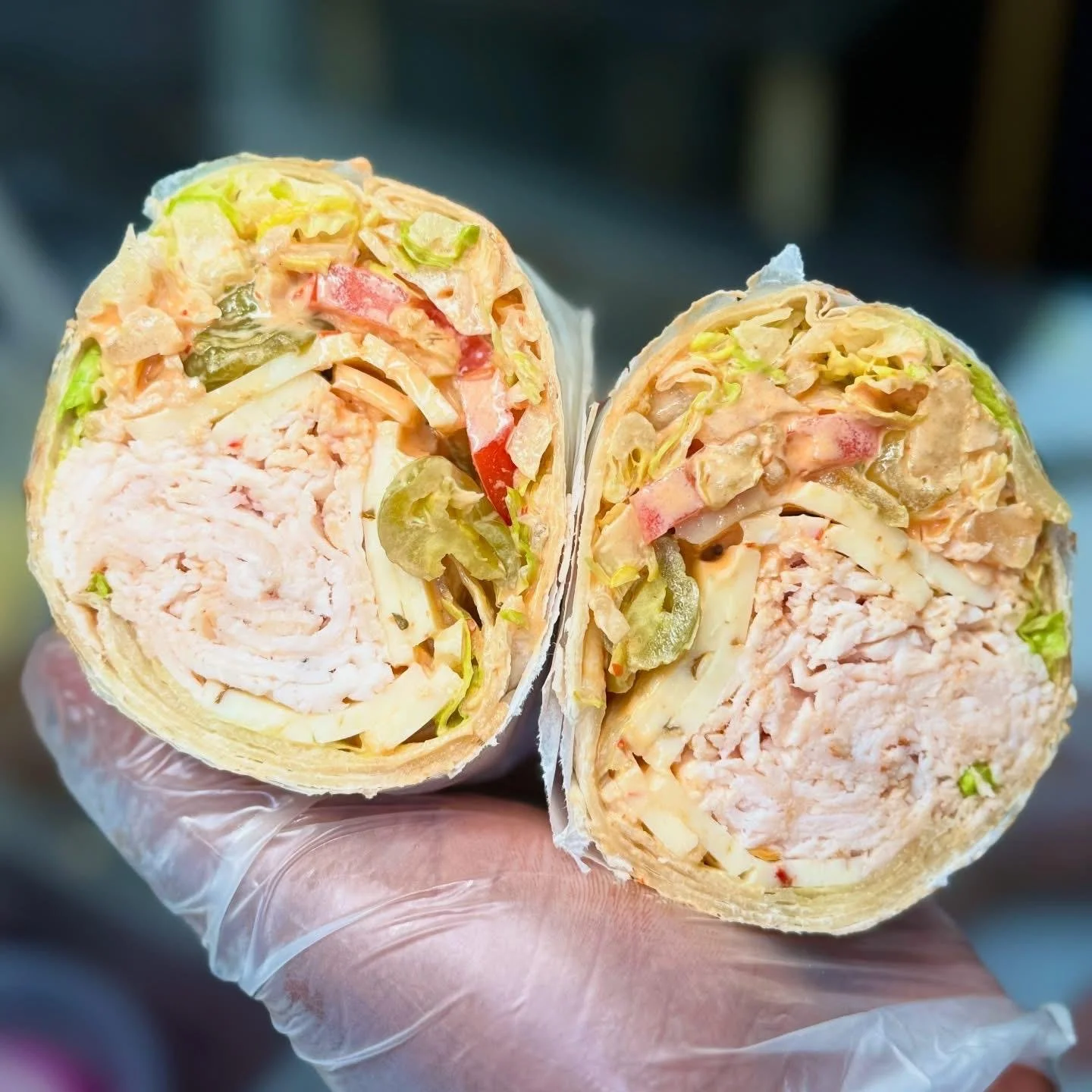 A hand wearing a disposable glove holding a sliced wrap sandwich filled with chicken, lettuce, tomatoes, pickles, and mayonnaise.