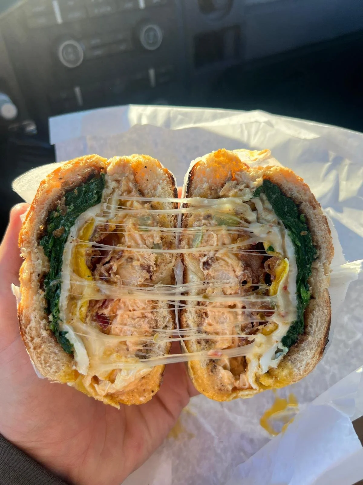 Close-up of a stuffed sandwich with melted cheese, spinach, chicken, and other fillings. The sandwich is cut in half and held by a hand inside a vehicle.