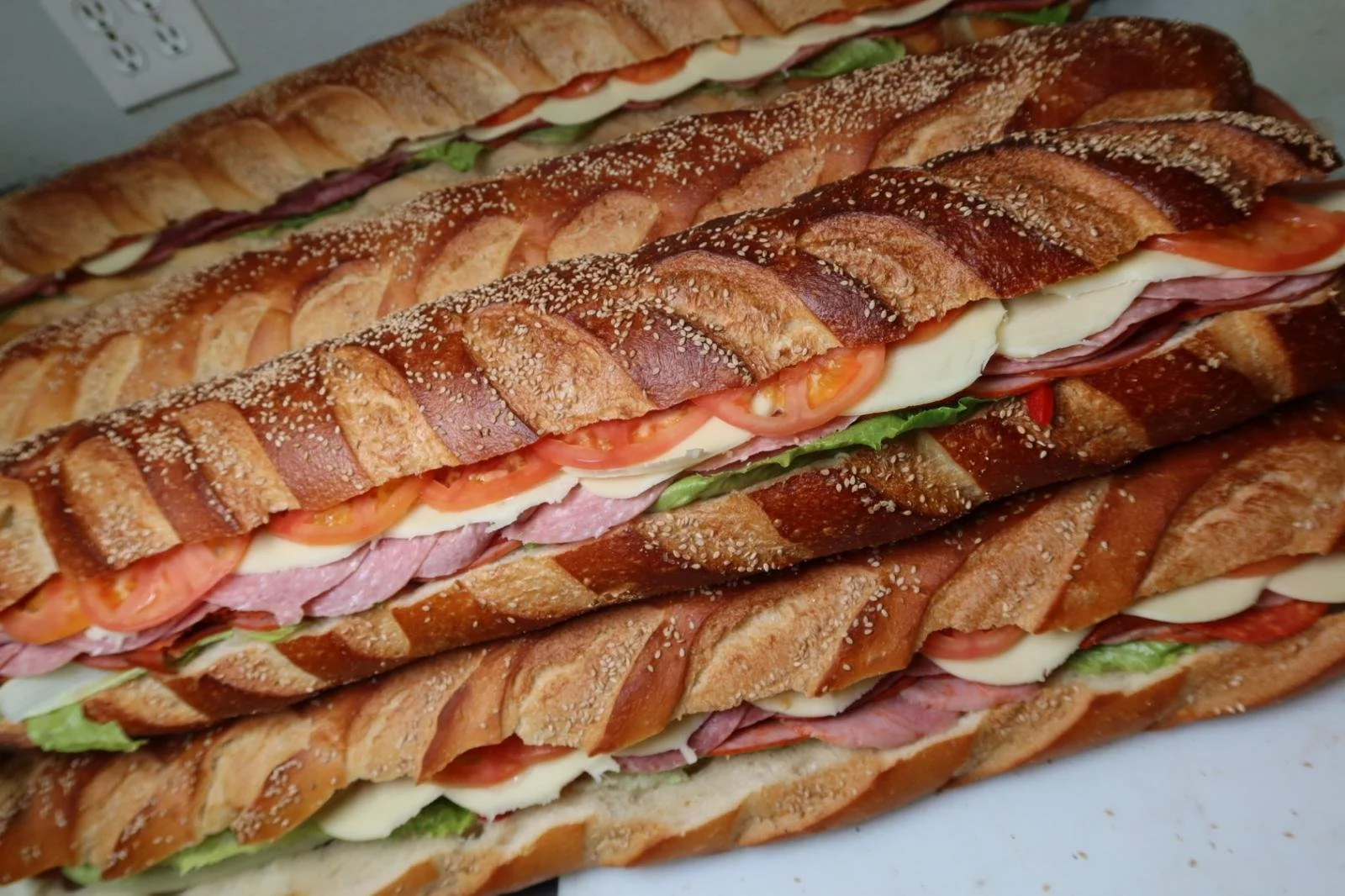 Large Italian hoagie sandwiches on a counter with layers of meats, cheese, tomatoes, lettuce, and condiments in subs with braided crusted bread.