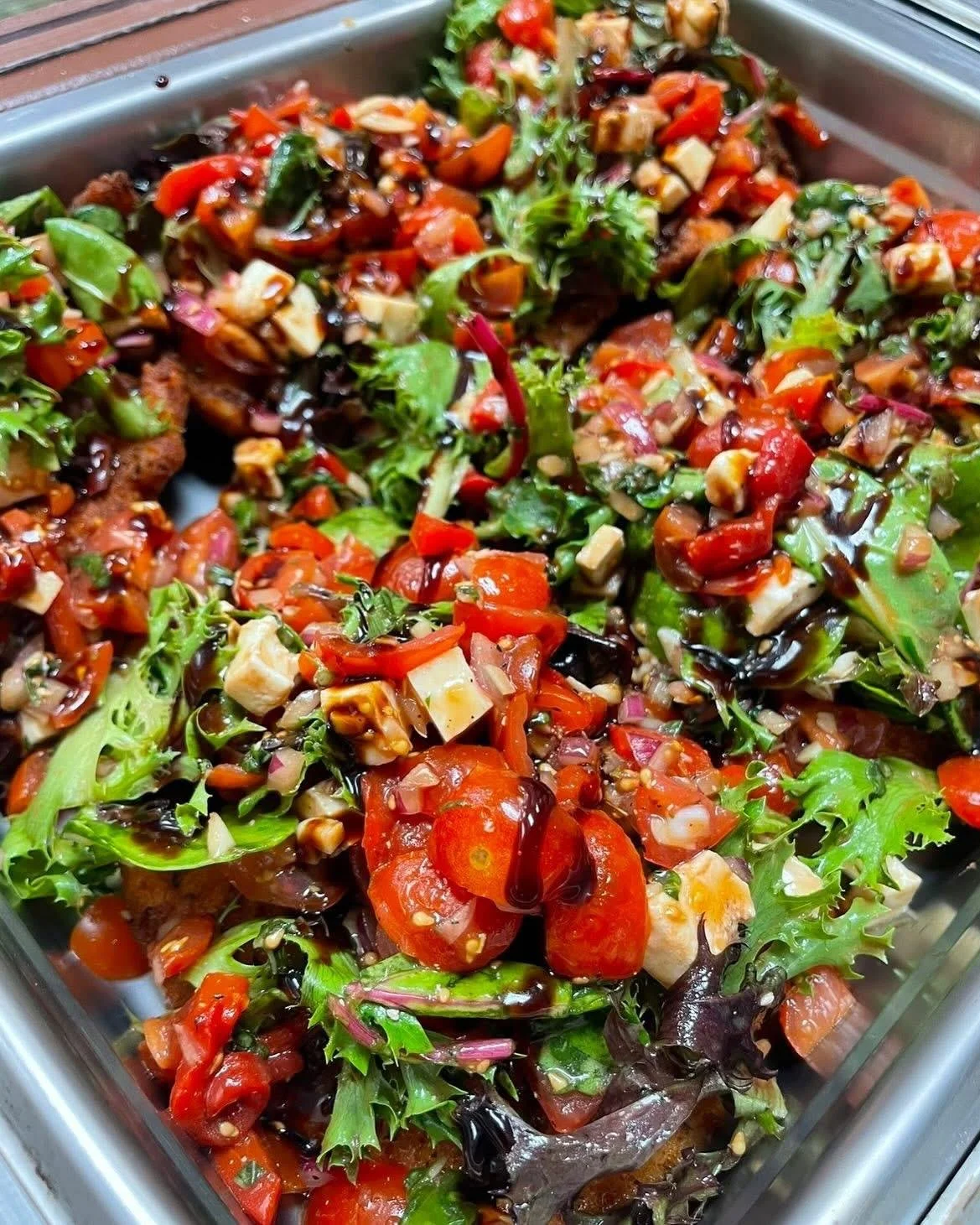 Mixed salad with cherry tomatoes, green leafy vegetables, chopped vegetables, nuts, and a dark drizzle dressing in a glass container.