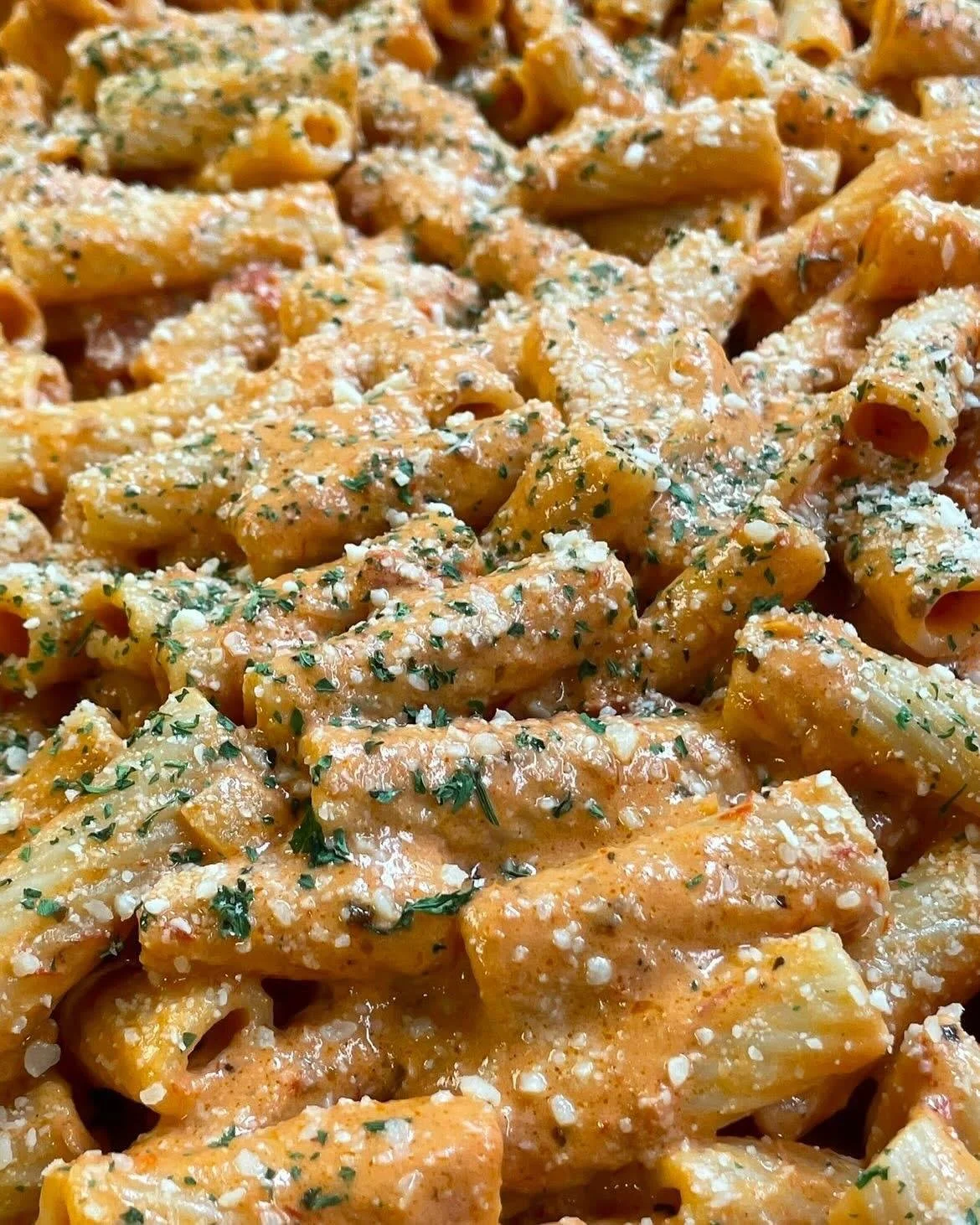 Close-up of cooked penne pasta coated in a creamy sauce with grated cheese and garnished with chopped herbs.