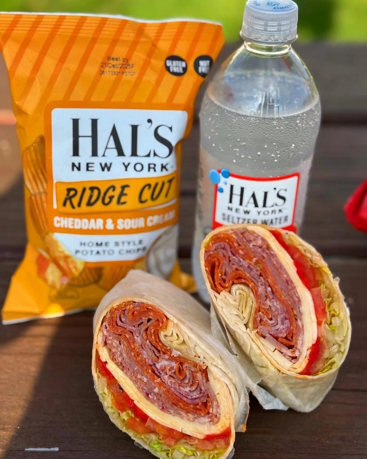 A wrap sandwich cut in half revealing layers of meat, cheese, and vegetables, placed on a wooden surface. In the background, there is a bag of Hal's New York Ridge Cut Cheddar & Sour Cream potato chips and a bottle of Hal's New York Seltzer Water.
