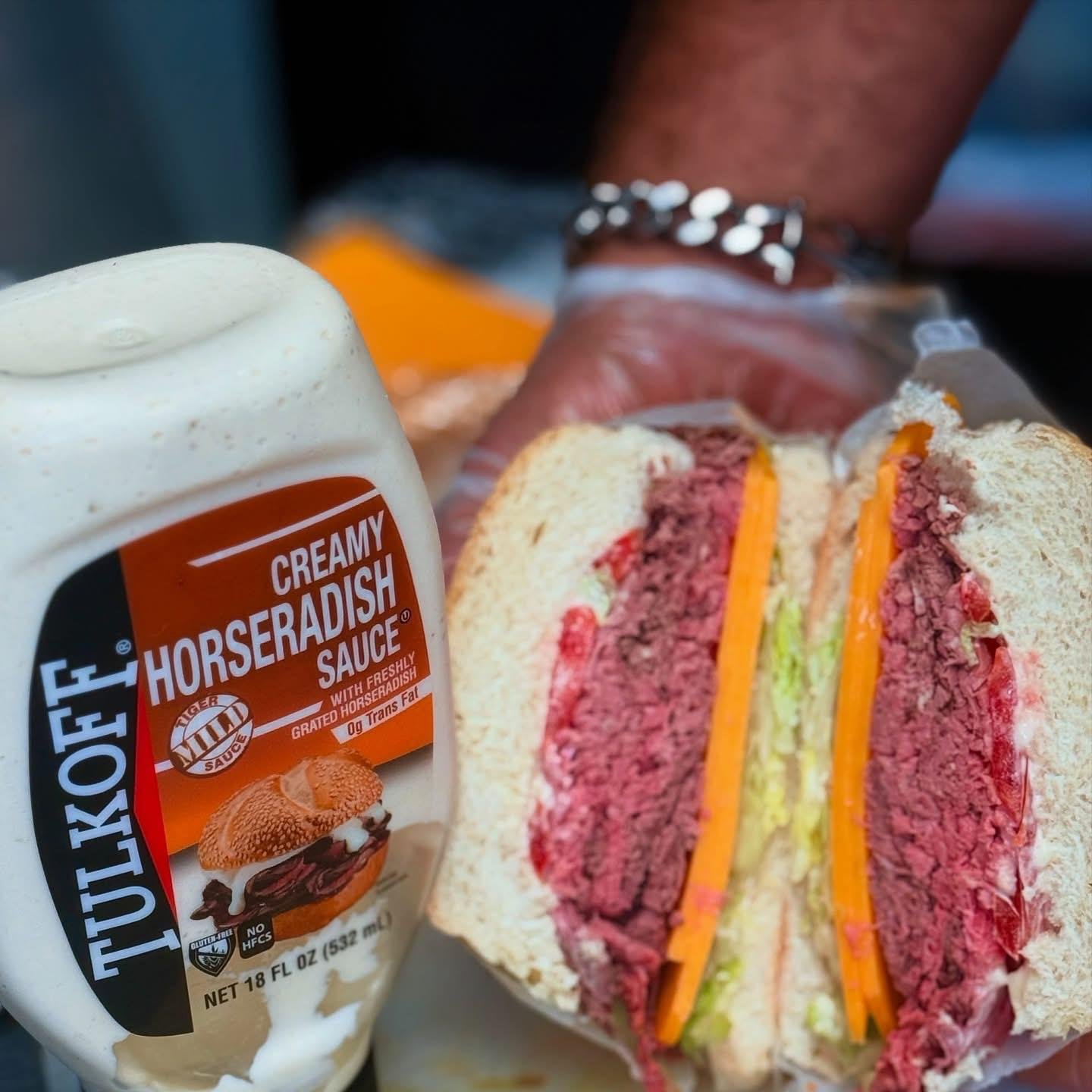 Close-up view of a sandwich with slices of roast beef, cheddar cheese, lettuce, and tomato between two halves of a sandwich bun. A container of creamy horseradish sauce with a label is placed beside the sandwich.