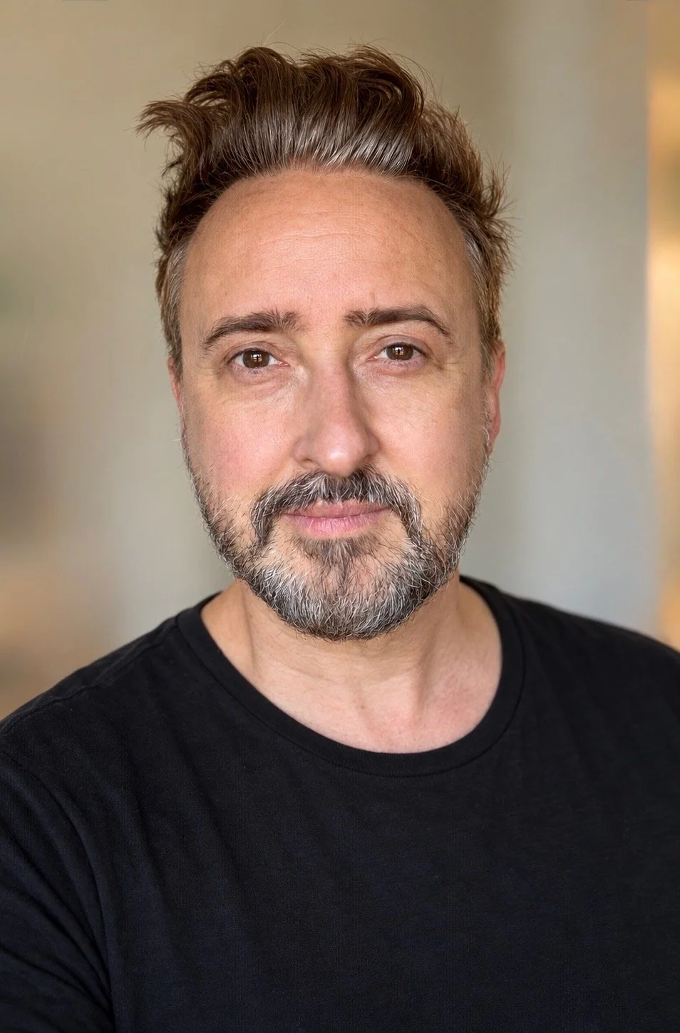 Close-up portrait of Tim Mattia the psychotherapist and therapeutic coach for the creative industries, wearing a black t-shirt, standing indoors with a blurred background.