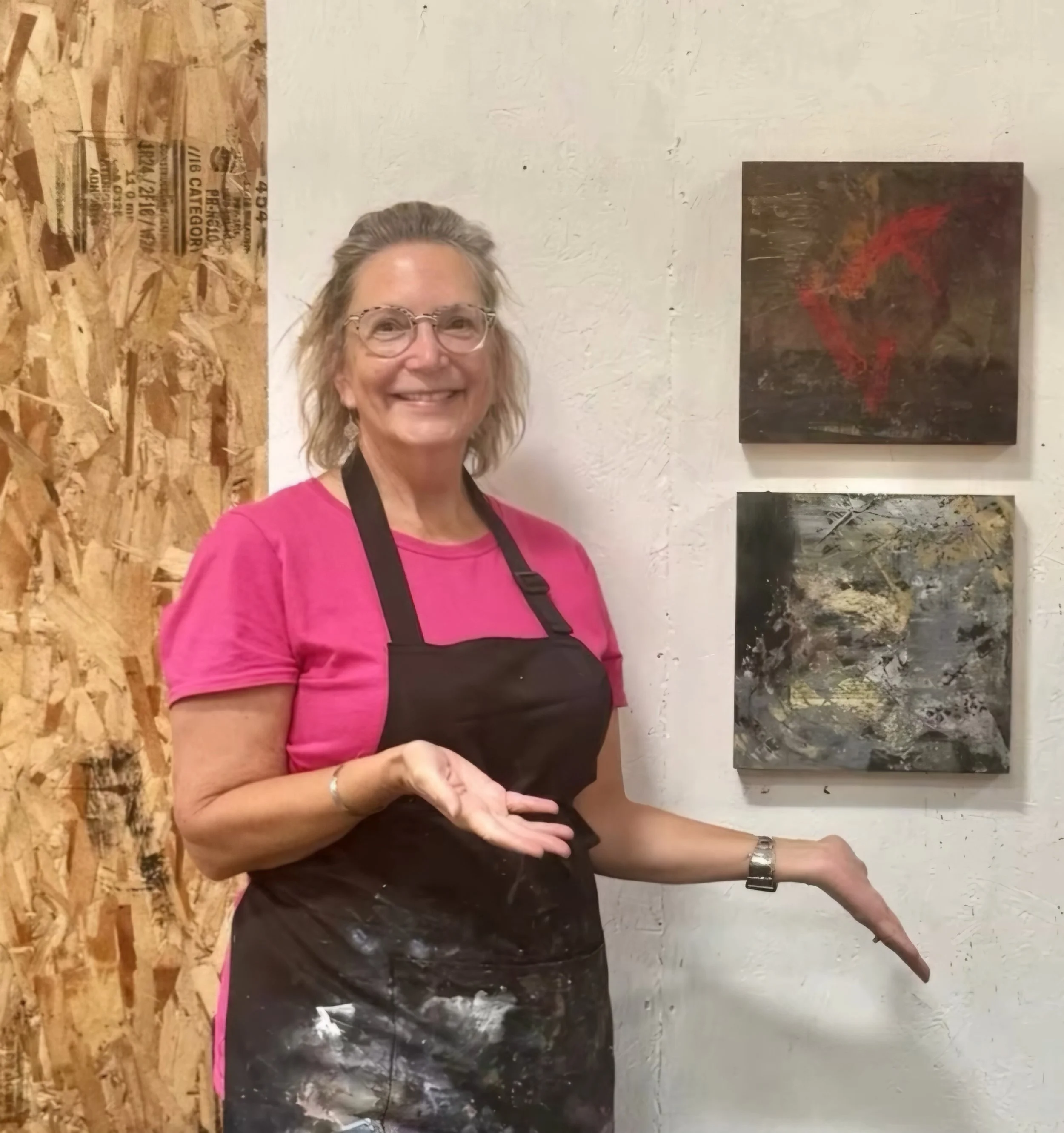 A woman with glasses, wearing a pink shirt and a black apron, smiling and standing in front of two abstract paintings on a white wall, with a wood panel on her left.