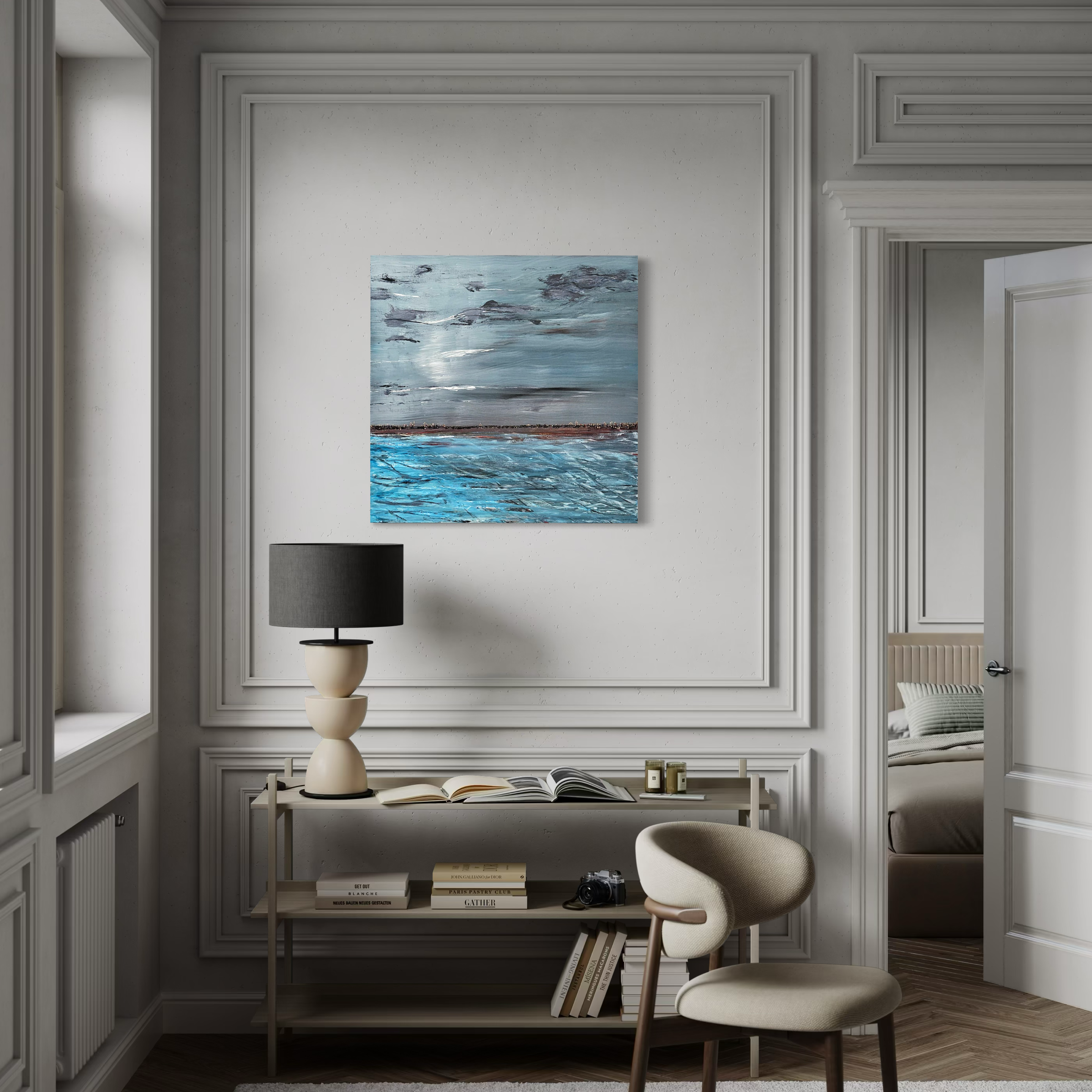 Interior of a modern room with white paneled walls, a small side table with books, a black lampshade, decorative candles, a camera, a beige armchair, and a landscape painting of sky, water, and distant land on the wall.