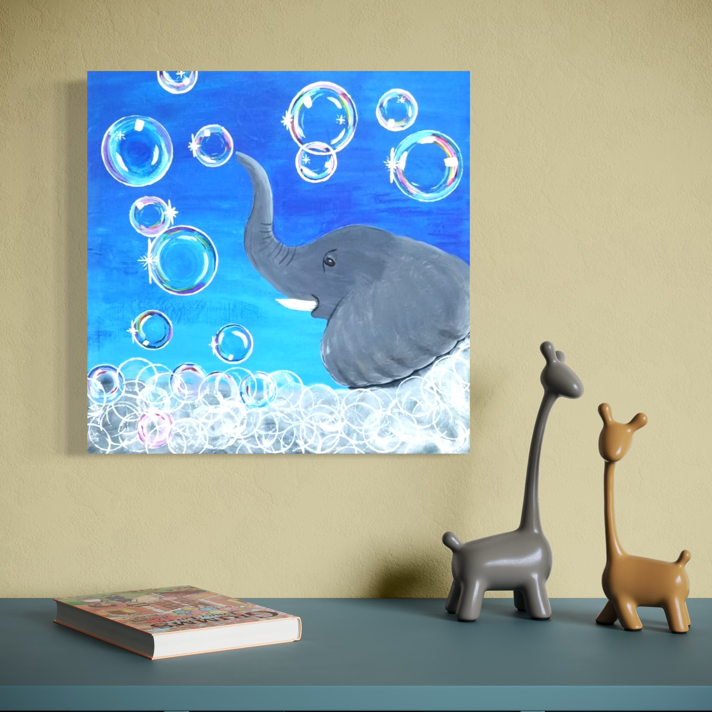 Children's art painting of an elephant with bubbles on a blue background, placed on a wall above a green table with two toy giraffes and a book.