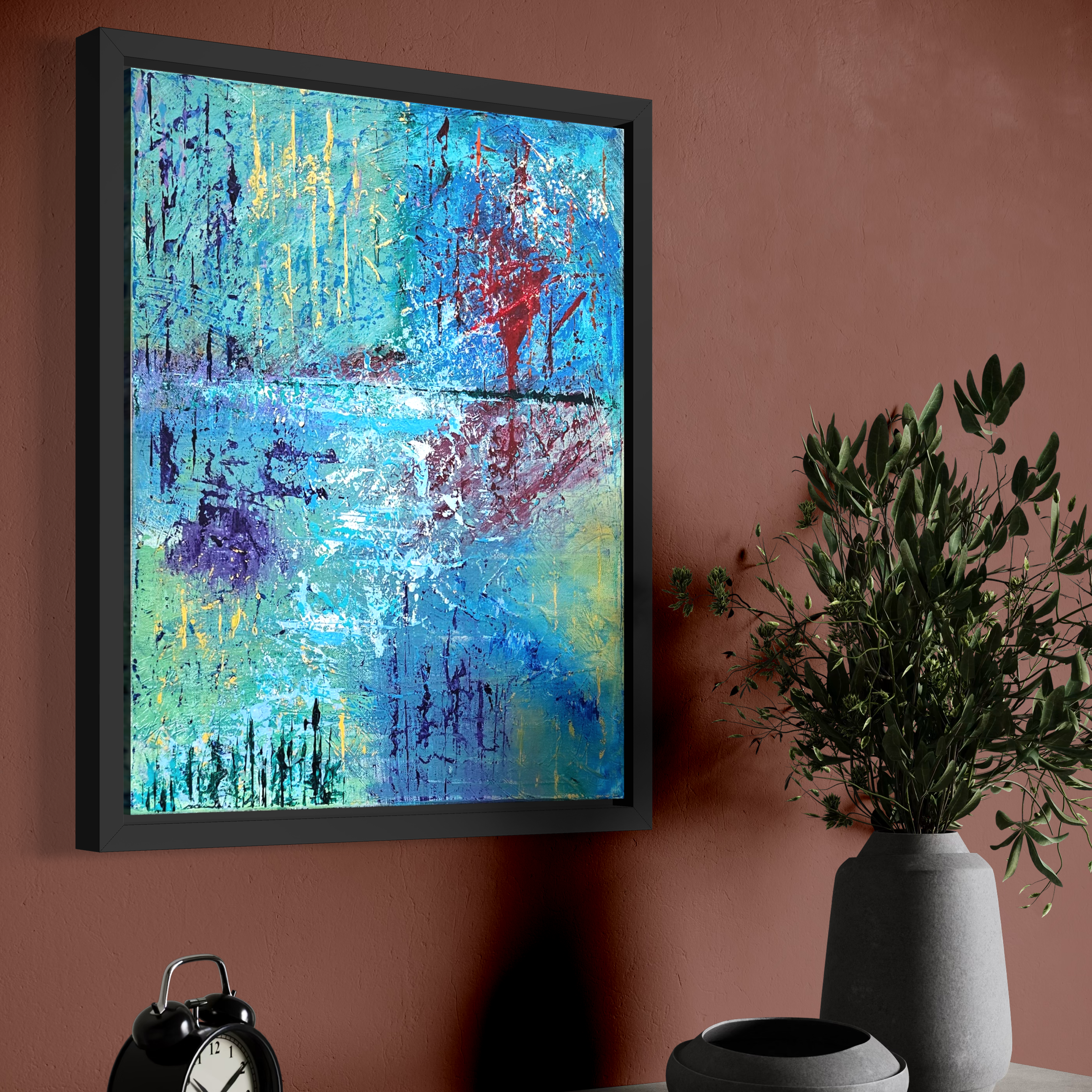 Abstract colorful painting with splashes of blue, yellow, purple, red, and white, framed and hung on a reddish-brown wall, with green plant in a gray vase and a silver clock on a surface below.