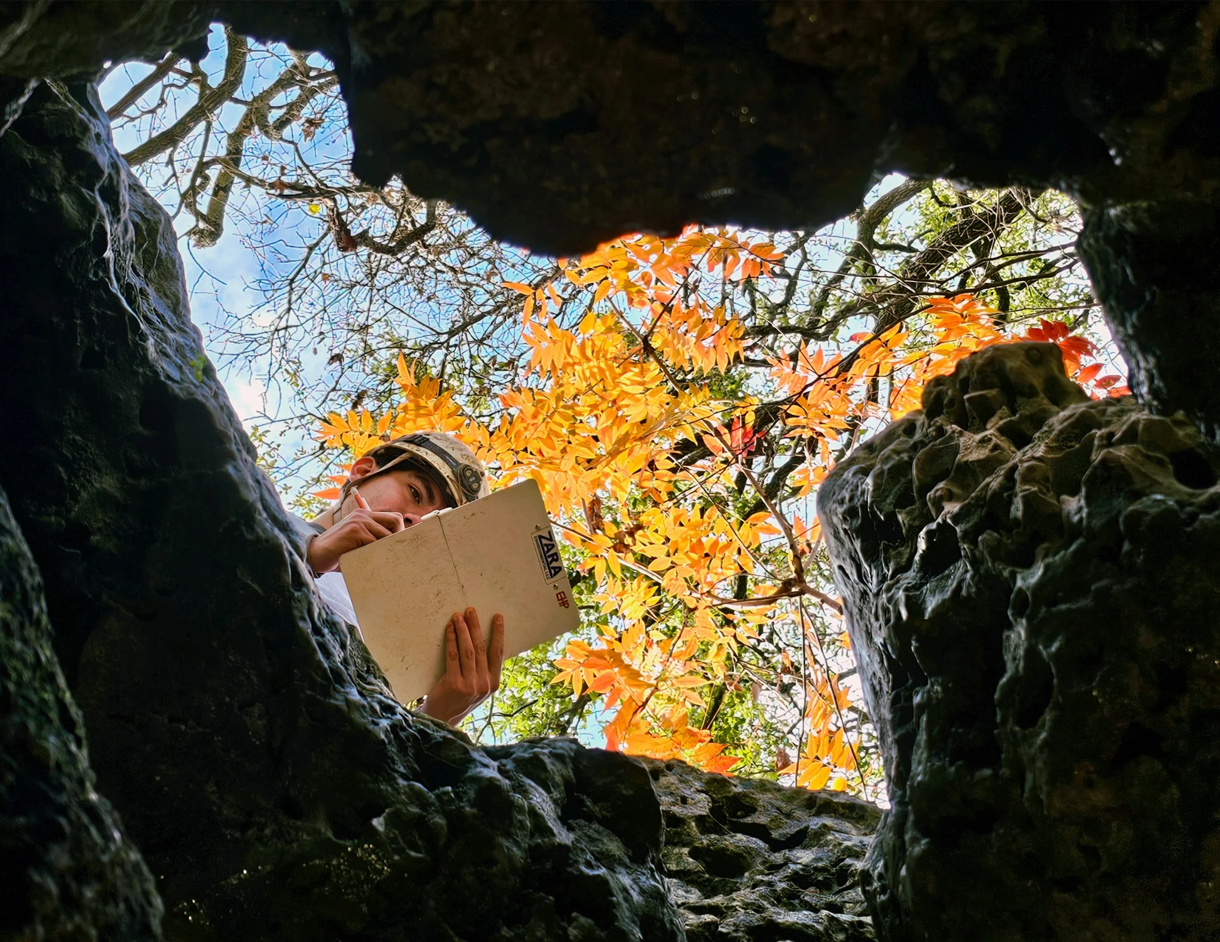 A person examining a rock formation from inside a hole in the ground, with autumn-colored trees and a blue sky visible above.