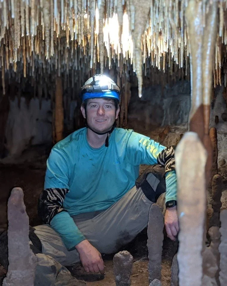 Man sitting inside a cave with stalactites hanging from the ceiling, wearing a helmet and outdoor clothing, smiling at the camera.