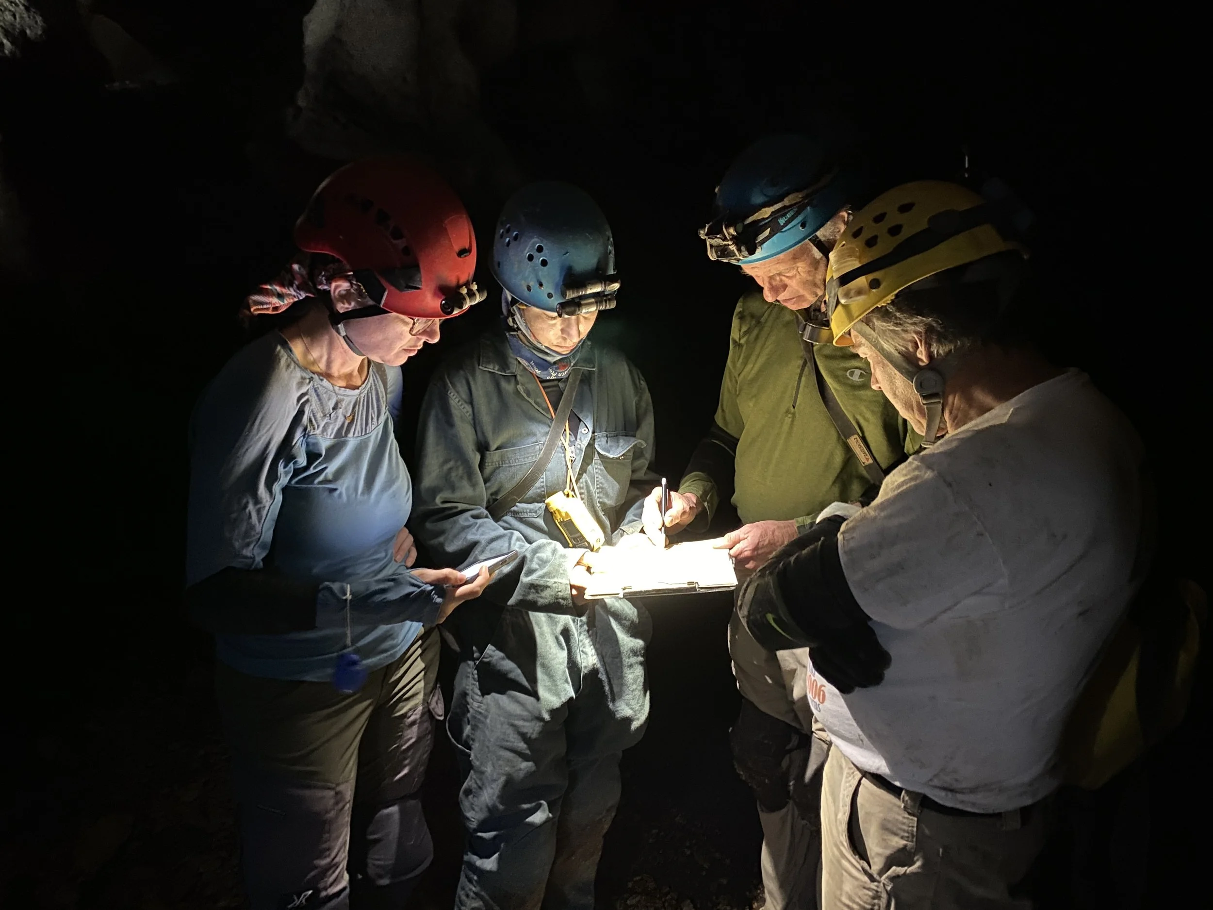 A group of five people wearing helmets and headlamps in a dark cave, gathered around a lit paper or notebook, engaged in a discussion or recording notes.