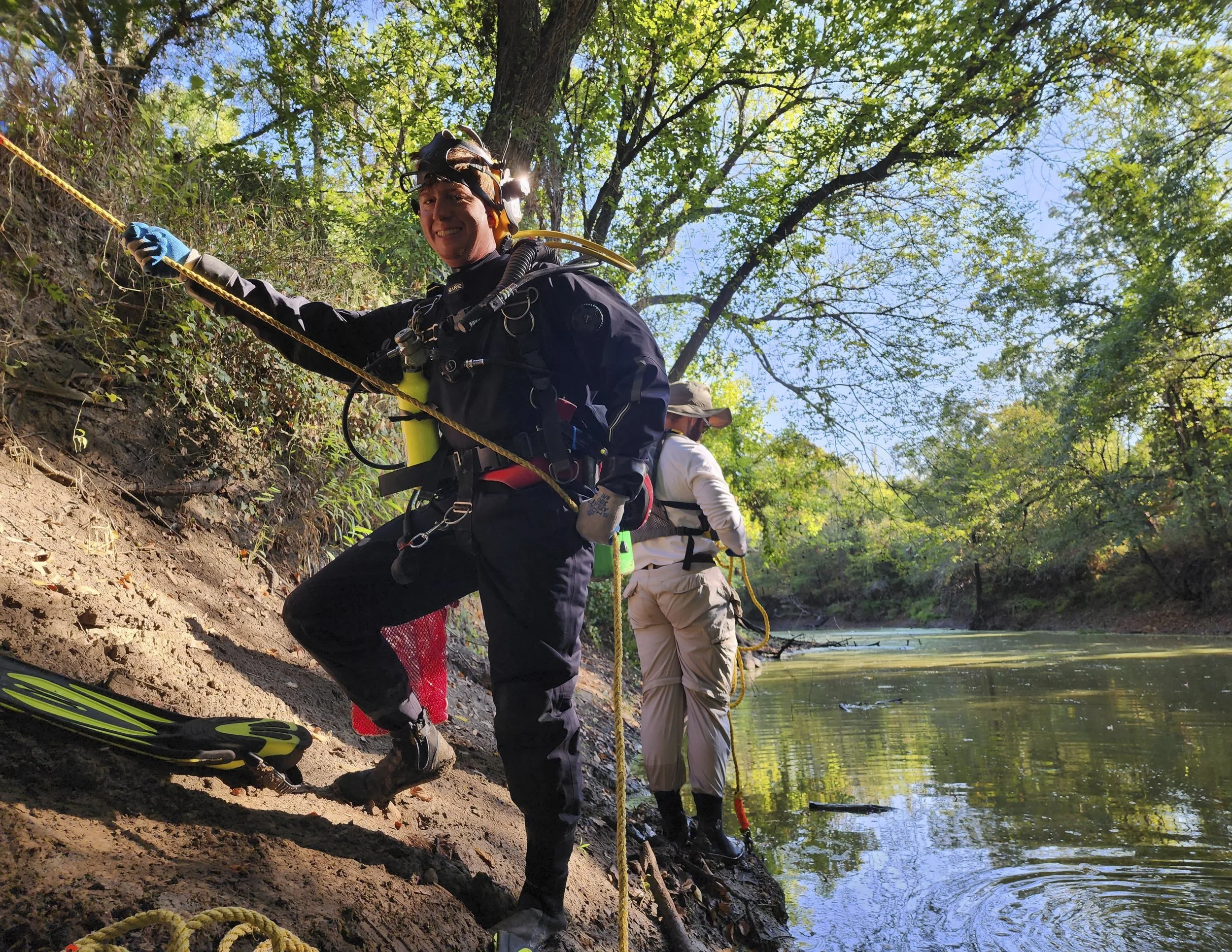 Two people in outdoor gear and helmets rappelling down a muddy riverbank surrounded by green trees.