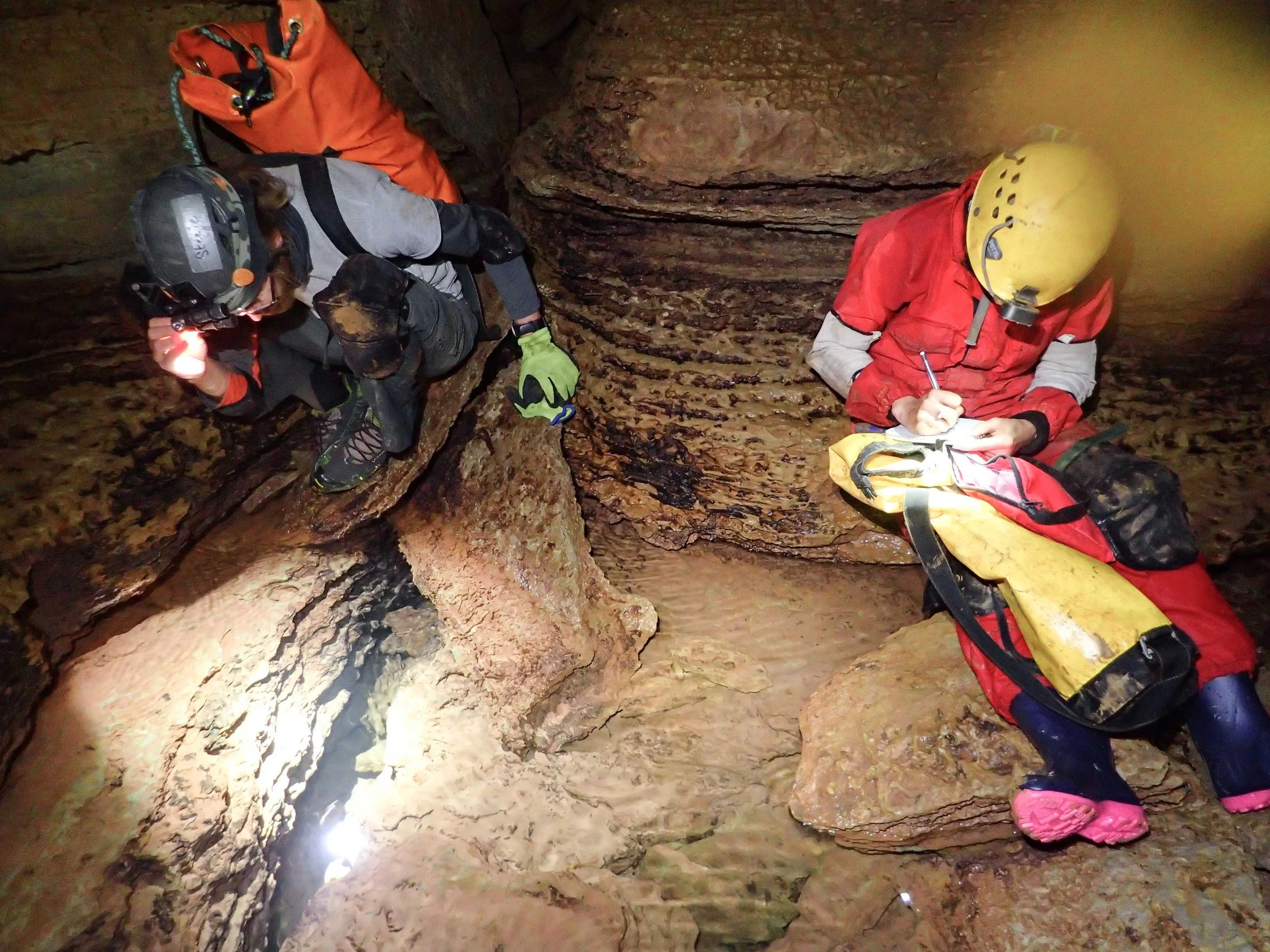 Two cavers inside a narrow, rocky cave. One is crouched, holding a flashlight, and the other is sitting on a rock, writing in a notebook.