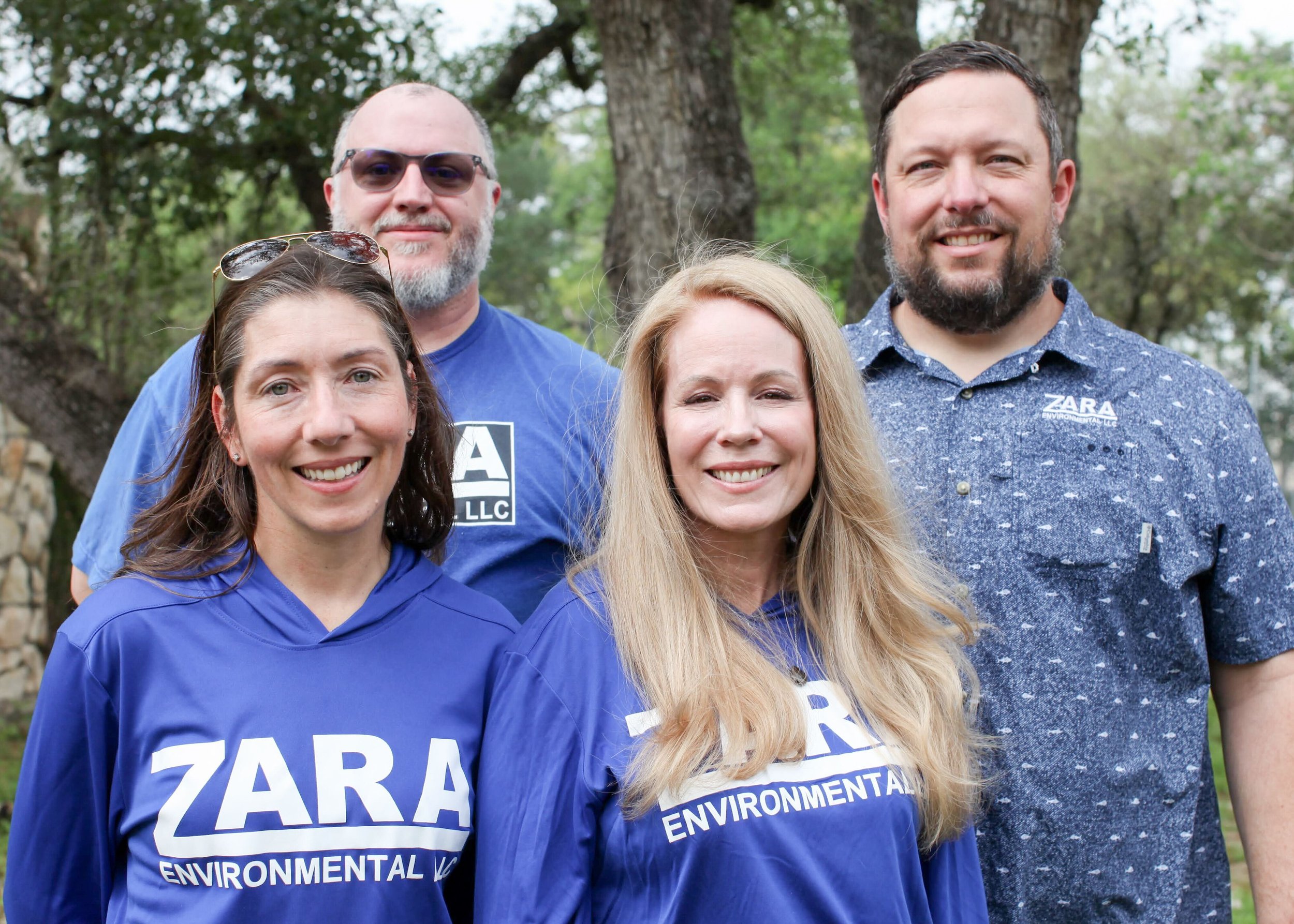 Five people standing outdoors in front of trees, smiling and facing the camera. Some are wearing blue shirts with 'ZARA Environmental' printed on them.
