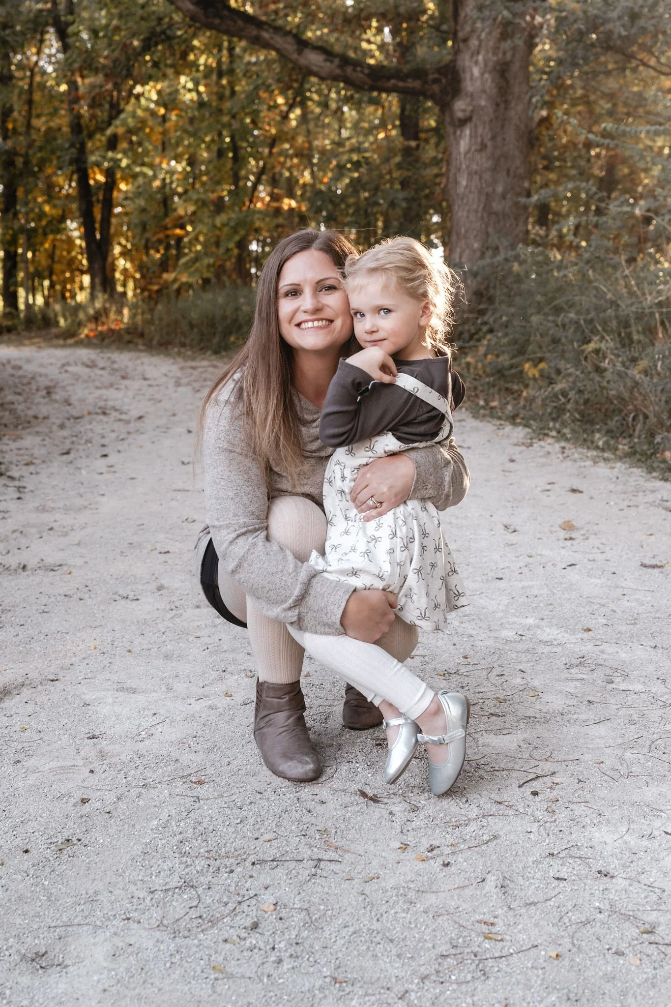 A woman and a young girl smiling and posing outdoors on a dirt path surrounded by trees with autumn foliage.