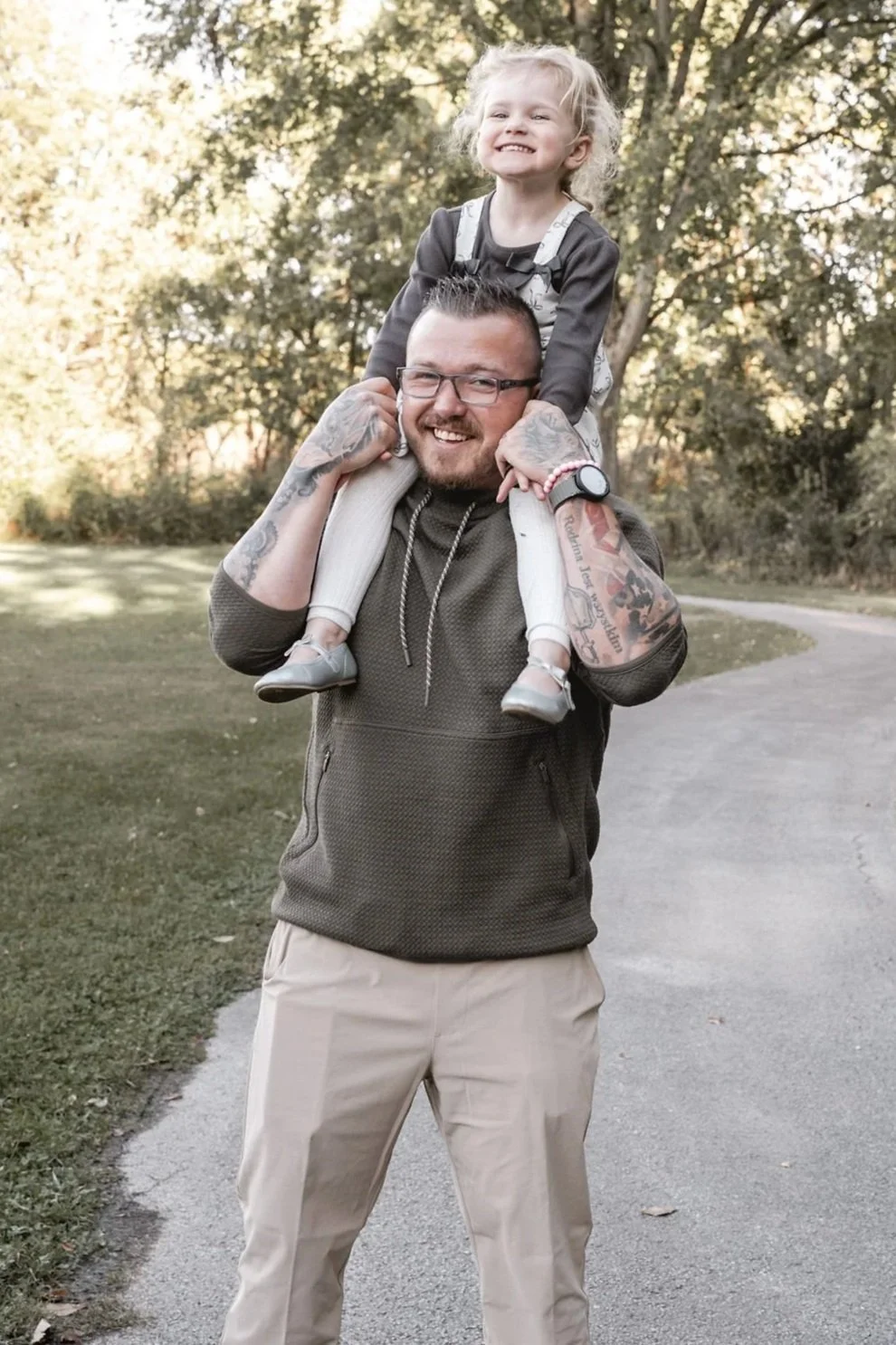 A man with tattoos and glasses carries a young girl on his shoulders outdoors on a park path during daytime. The girl is smiling and gripping his head.