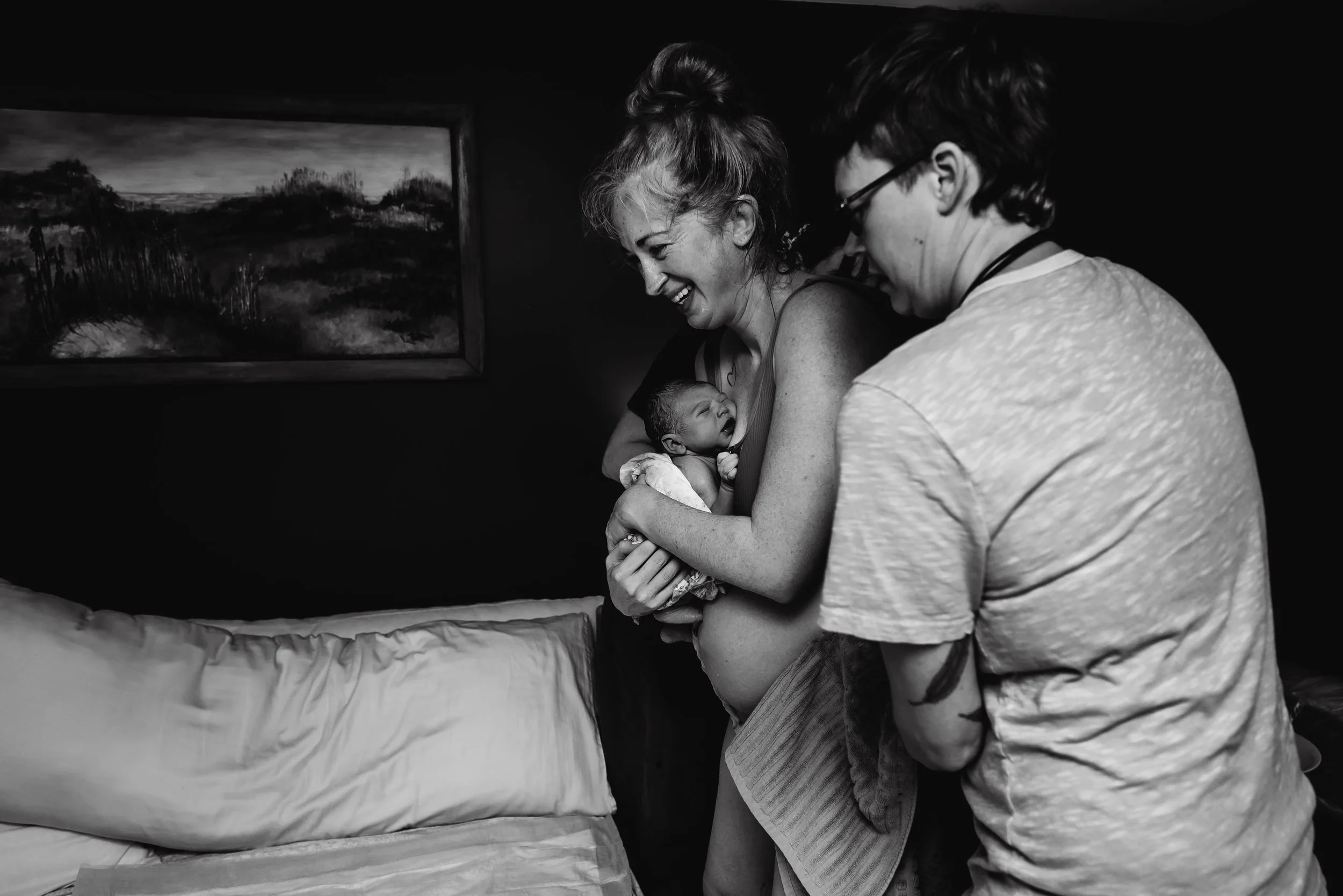 A woman is holding a newborn baby, smiling and sharing a joyful moment with a midwife standing next to her, in a room with dark walls and a landscape painting.