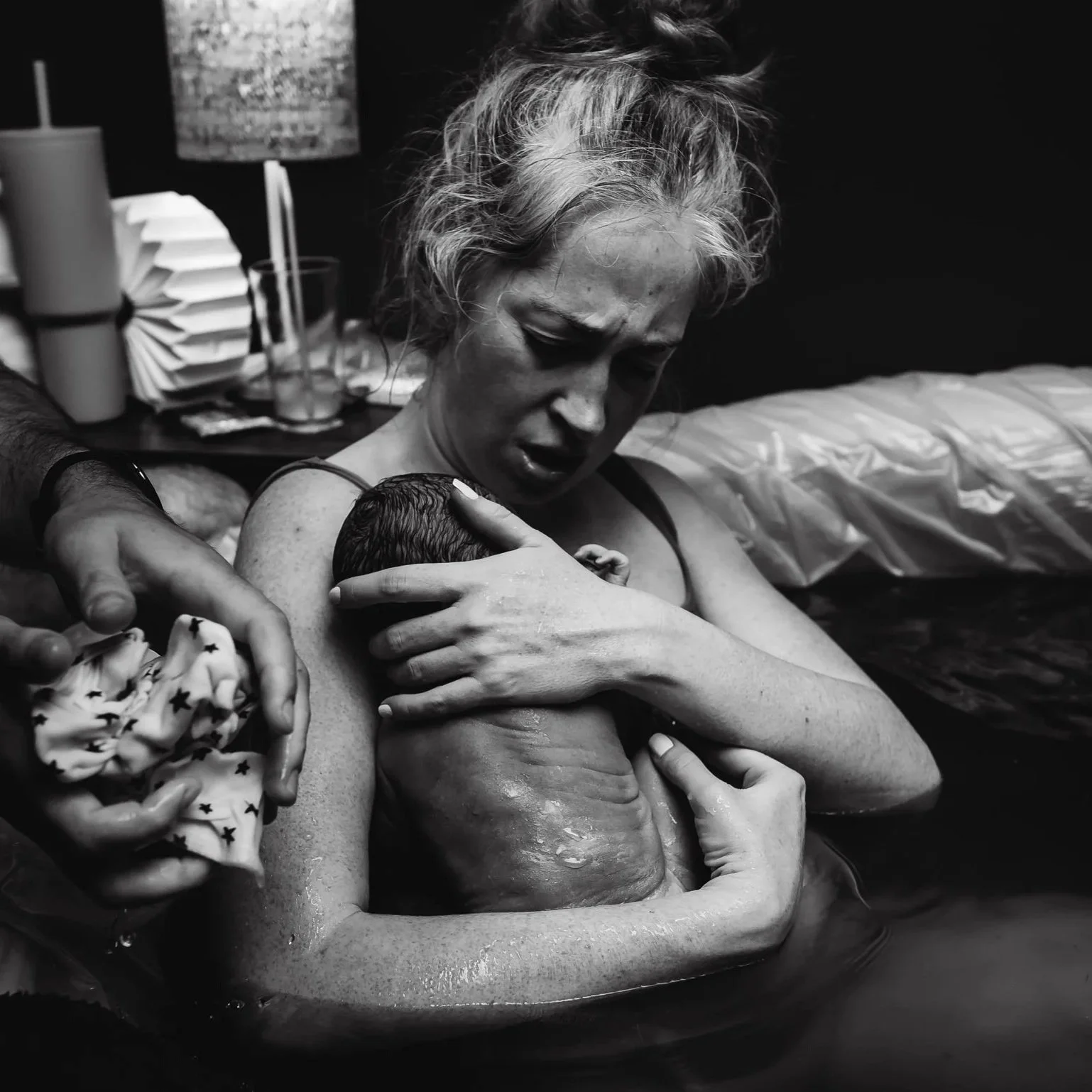 A woman holding a newborn baby during home childbirth, with a look of relief and emotion, in a dimly lit room.