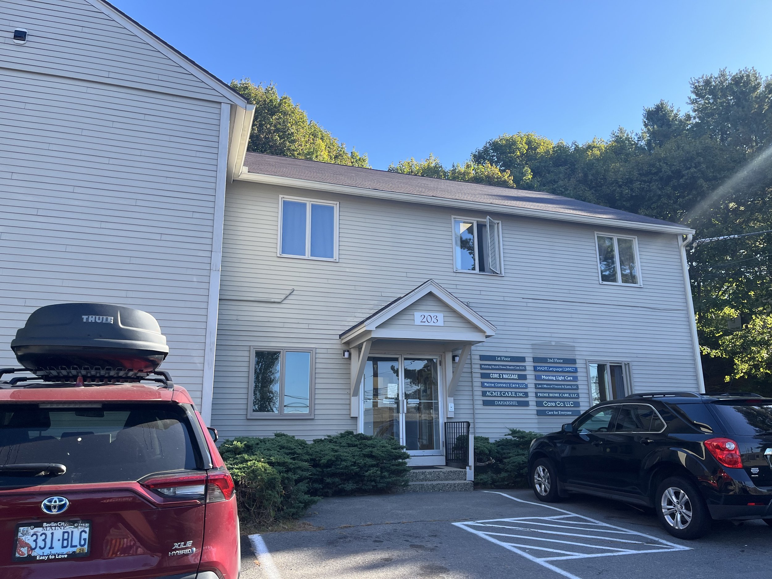 A two-story white building with signs on the right side, parking spaces in front, a red SUV with a roof cargo box on the left, and a black SUV on the right, with trees and a blue sky in the background.