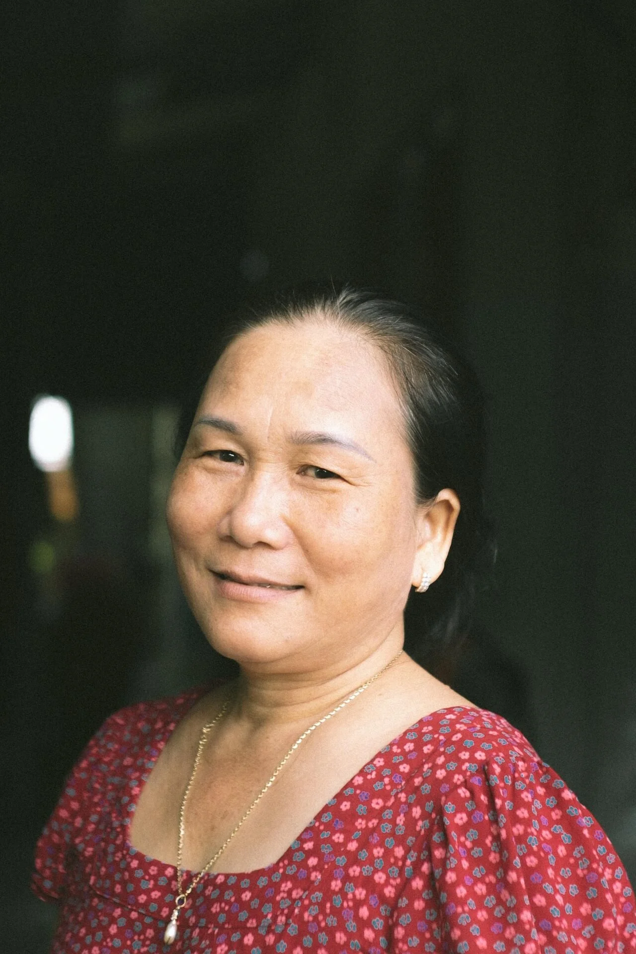 A smiling woman with tan skin and black hair styled back, wearing a red dress with a floral pattern, gold jewelry, and earrings against a dark background.