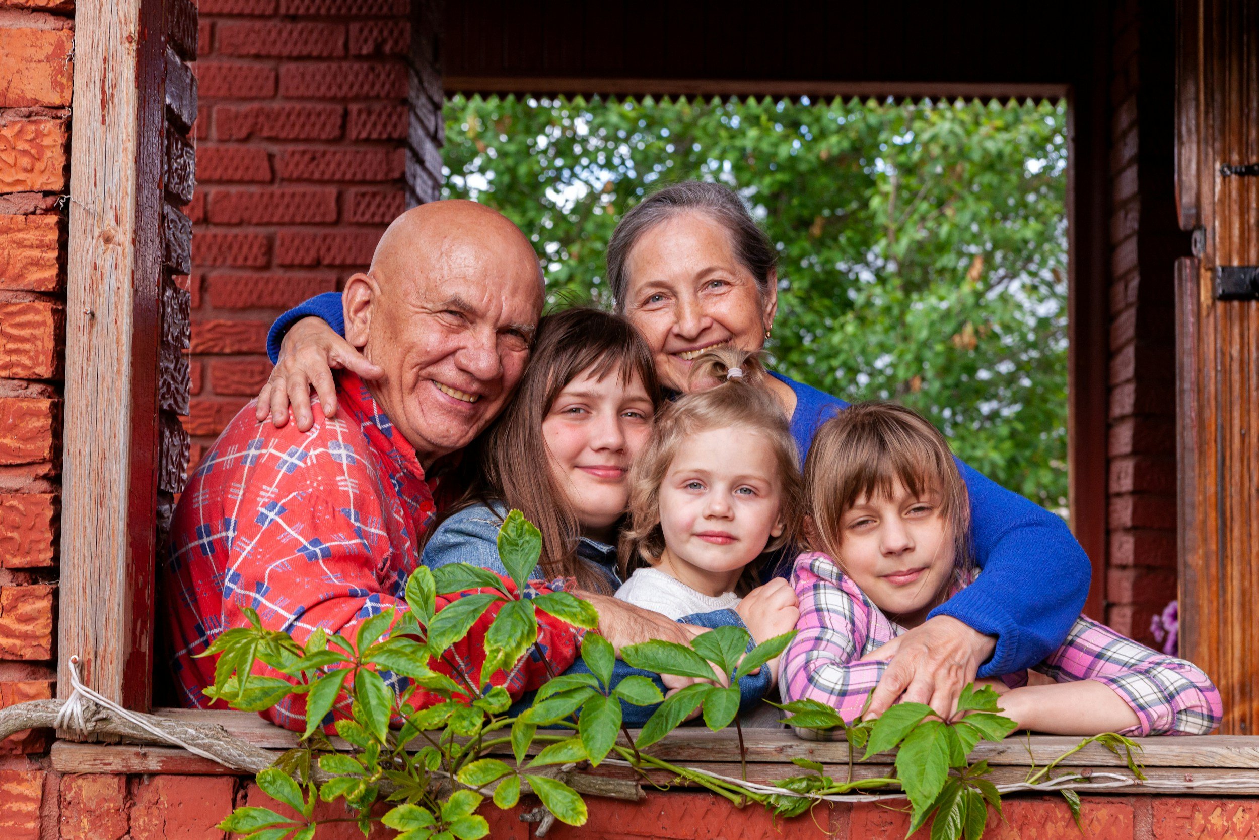 A multigenerational family of six smiling and hugging at a window of a red brick house, with green trees in the background.