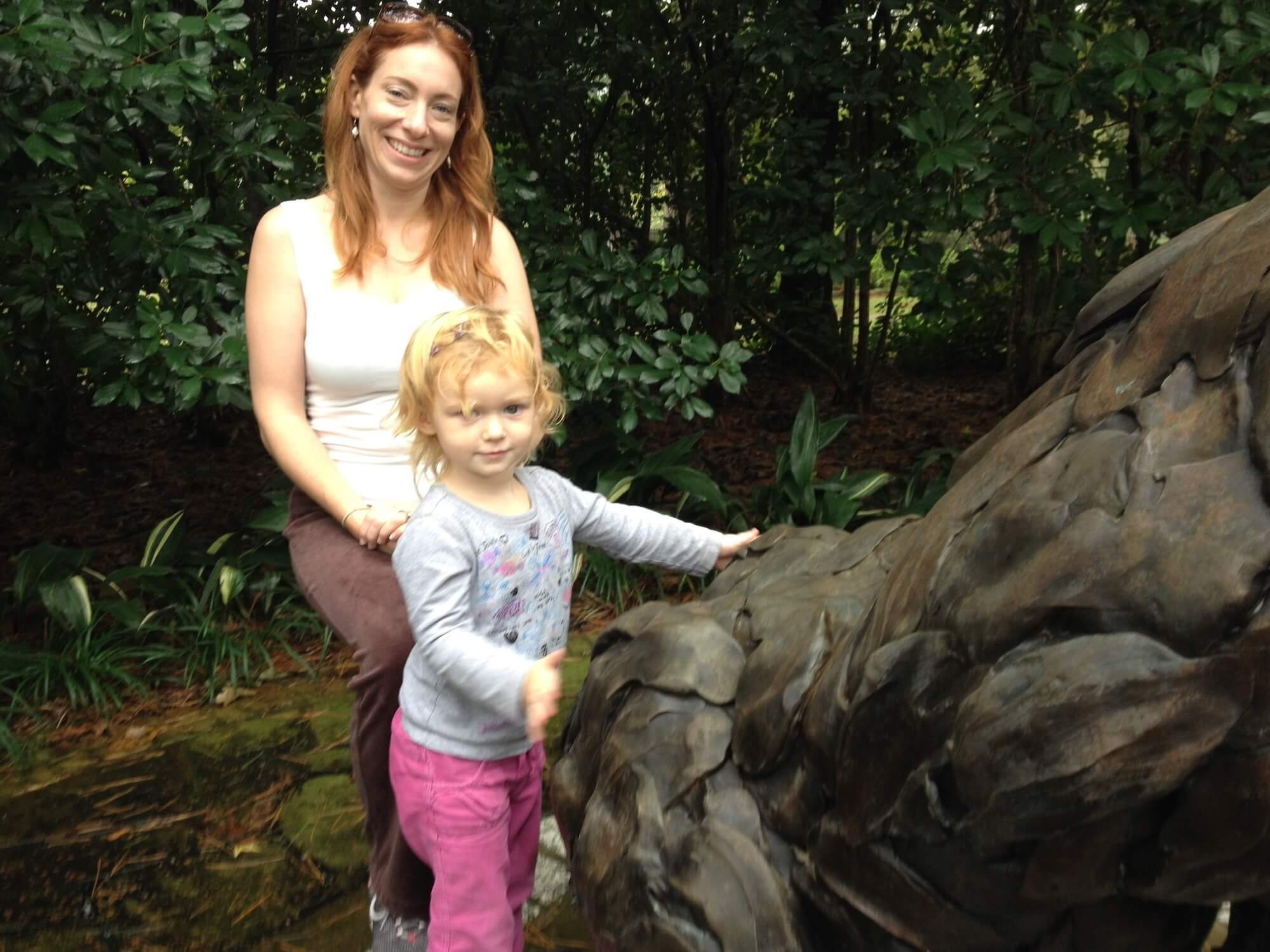 A woman and a young girl standing in a garden next to a large, textured rock, with greenery in the background.