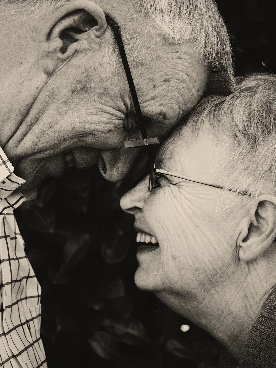 Two elderly individuals, a man and a woman, are smiling and touching foreheads in a close, affectionate moment.