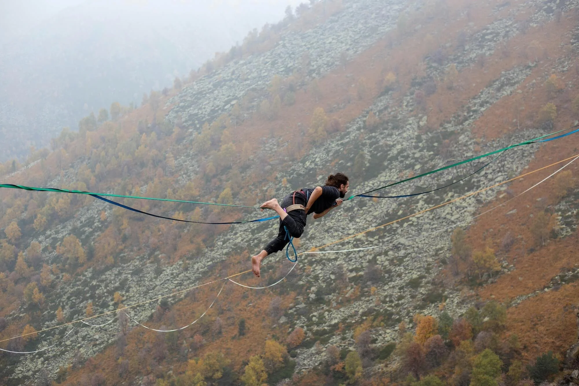 A person with long hair is walking on a tightrope high above a hillside covered in autumn-colored trees, during foggy weather.