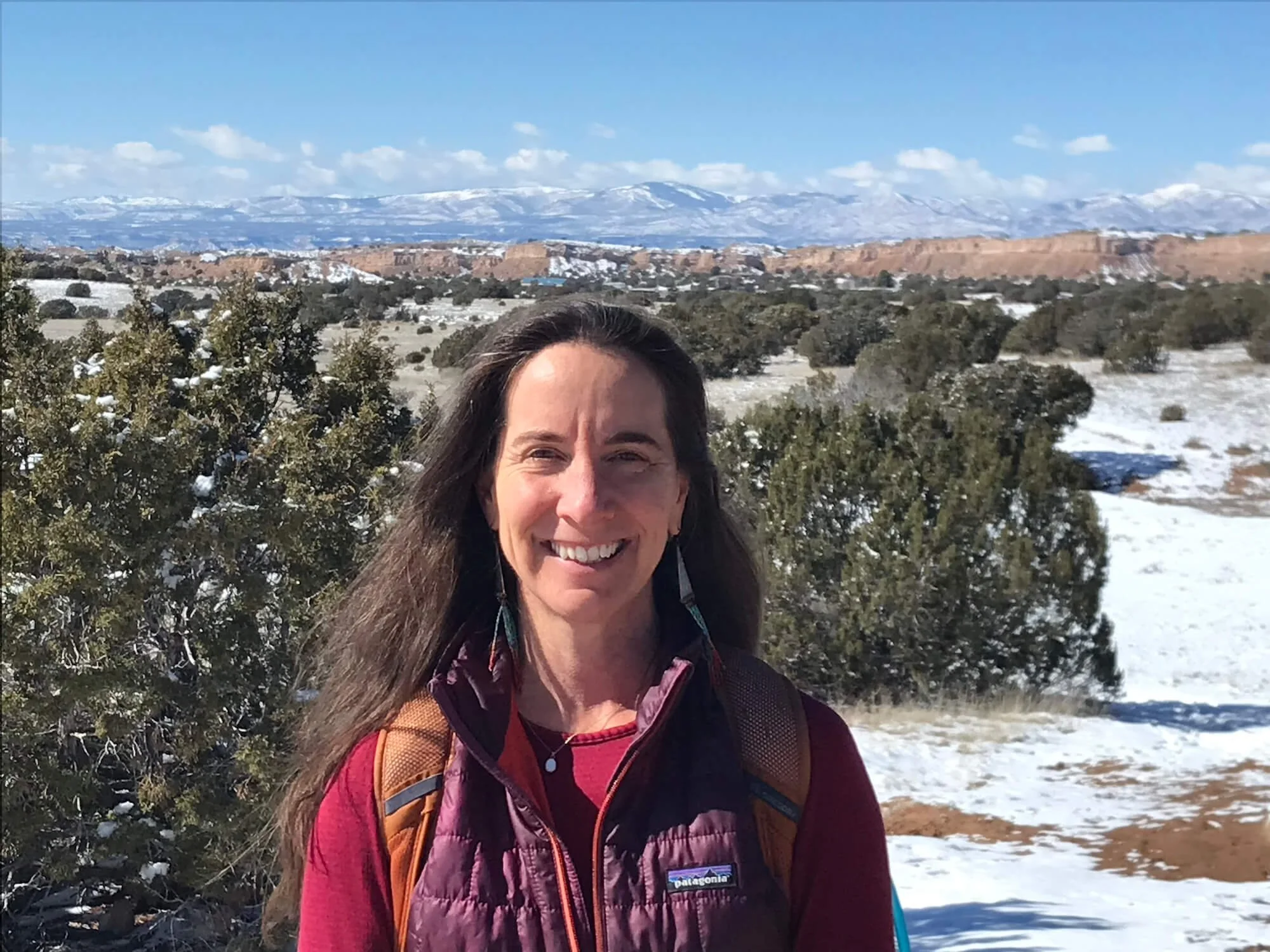 A woman with long brown hair taking a selfie outdoors in a snowy mountainous landscape. She is wearing a maroon jacket and a backpack.